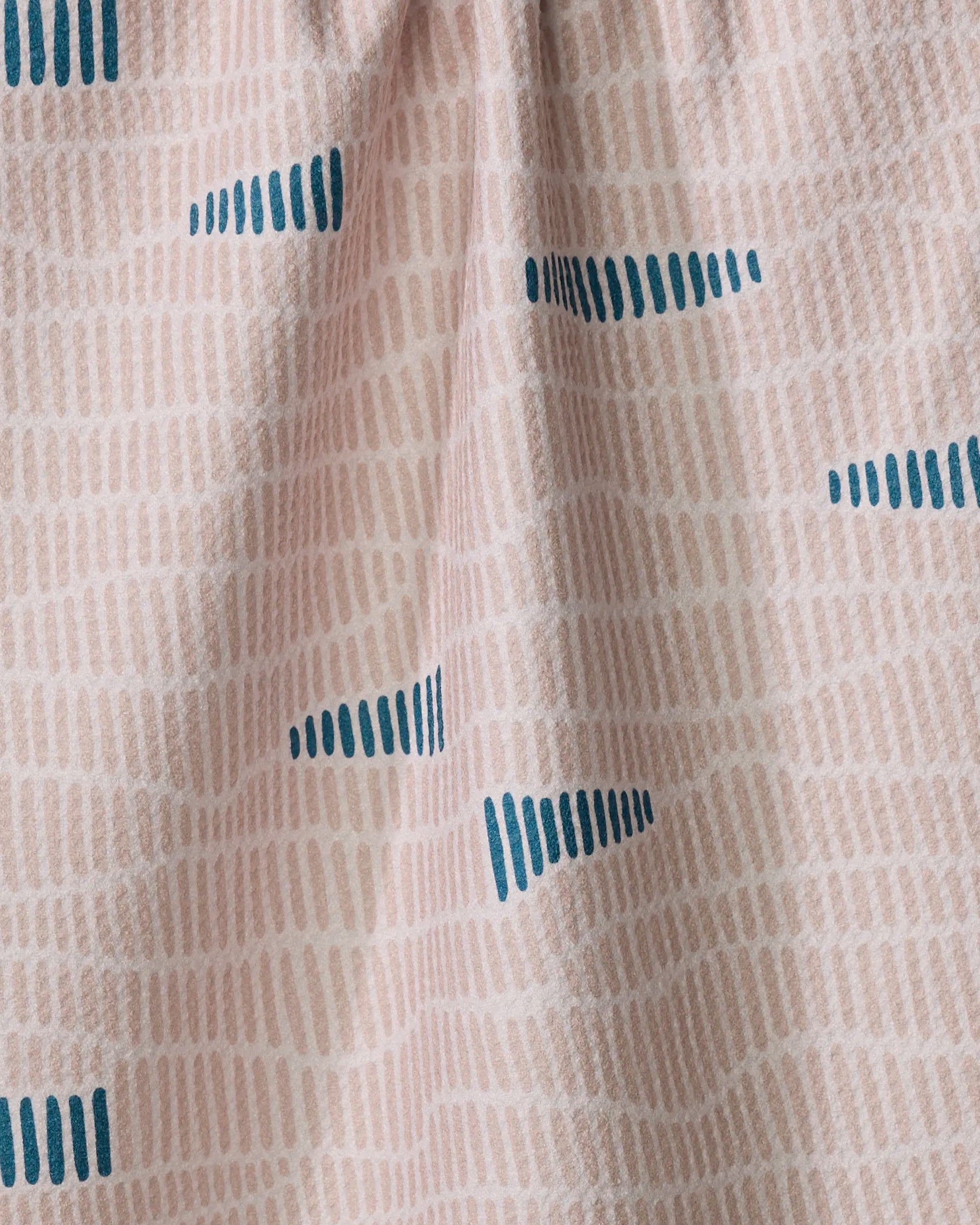 The Ridgeline tea towel by Twig + Spark features a light pink color, ultra-absorbent fabric, a soft beige grid pattern with scattered dark blue dashes, and a gently draped texture that adds visual interest.