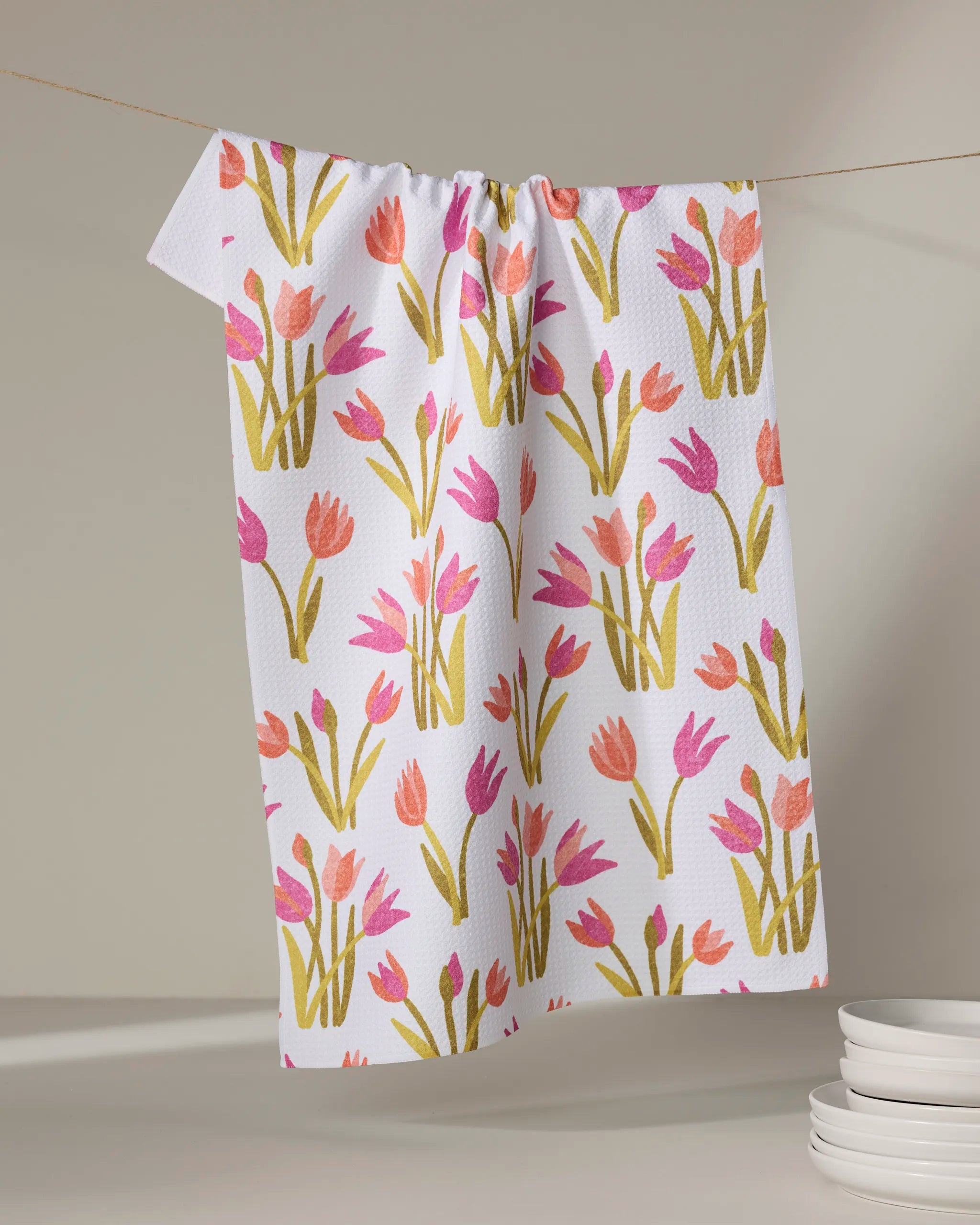 The Riley kitchen towel by Amy MacCready, featuring pink and orange tulips, hangs on a line with a small stack of white plates in the lower right corner.