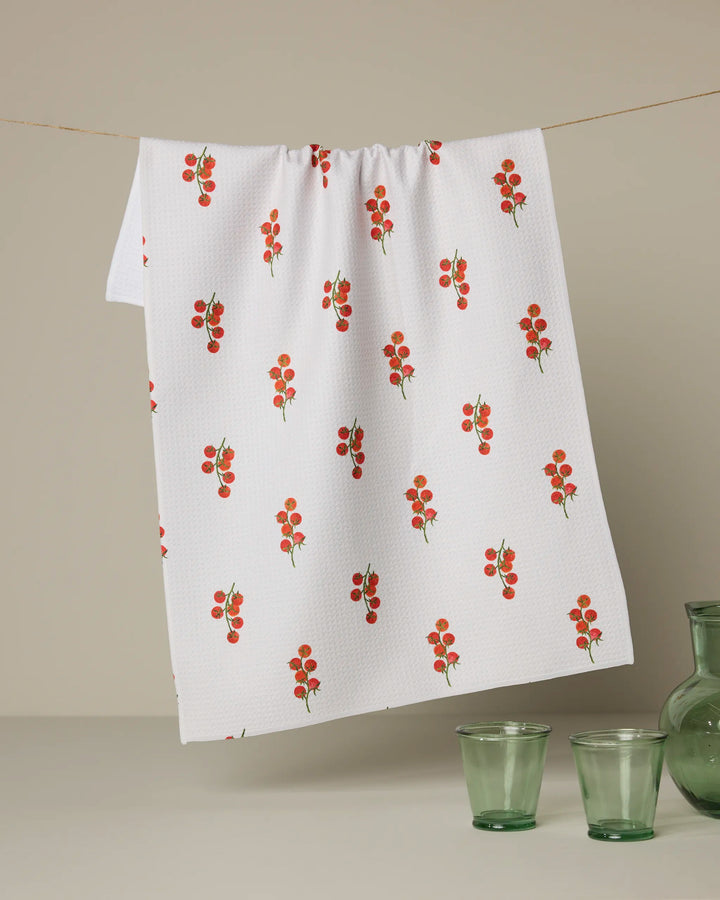 The Geometry "Ripe and Ready Scattered" recycled kitchen towel, featuring small red cherry tomato clusters, hangs on a line above two green glass cups and a matching vase.