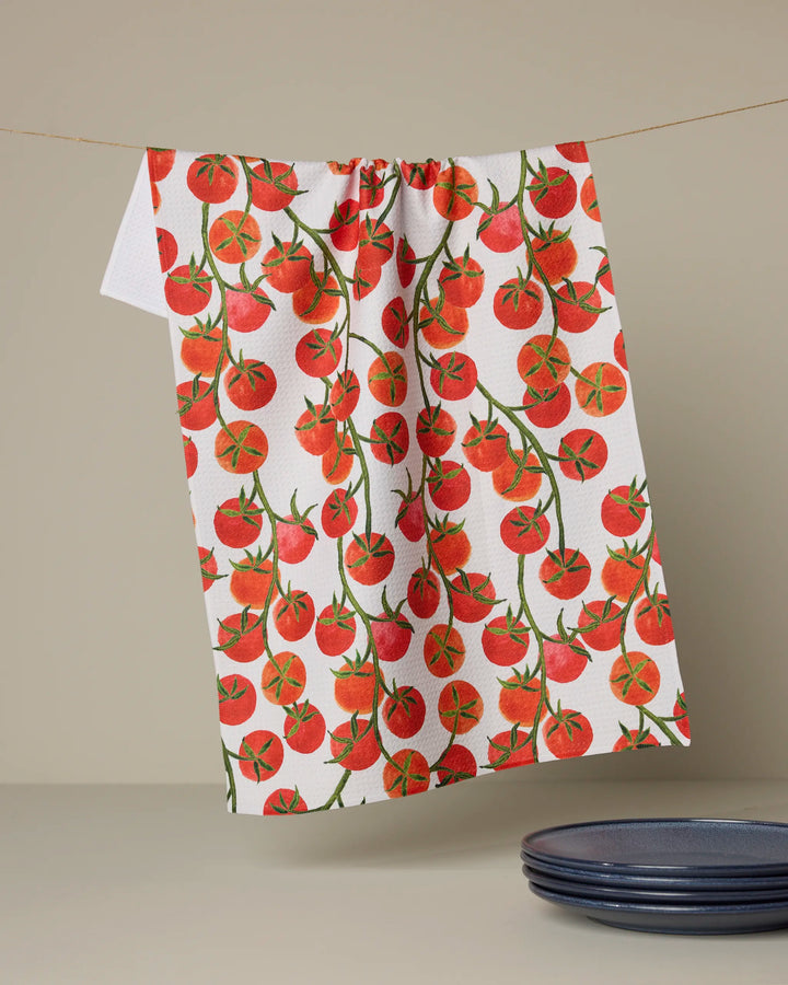 The Geometry "Ripe and Ready" tea towel, an eco-friendly white towel featuring vibrant red tomatoes and green stems, hangs above a neutral surface beside a stack of dark blue plates.