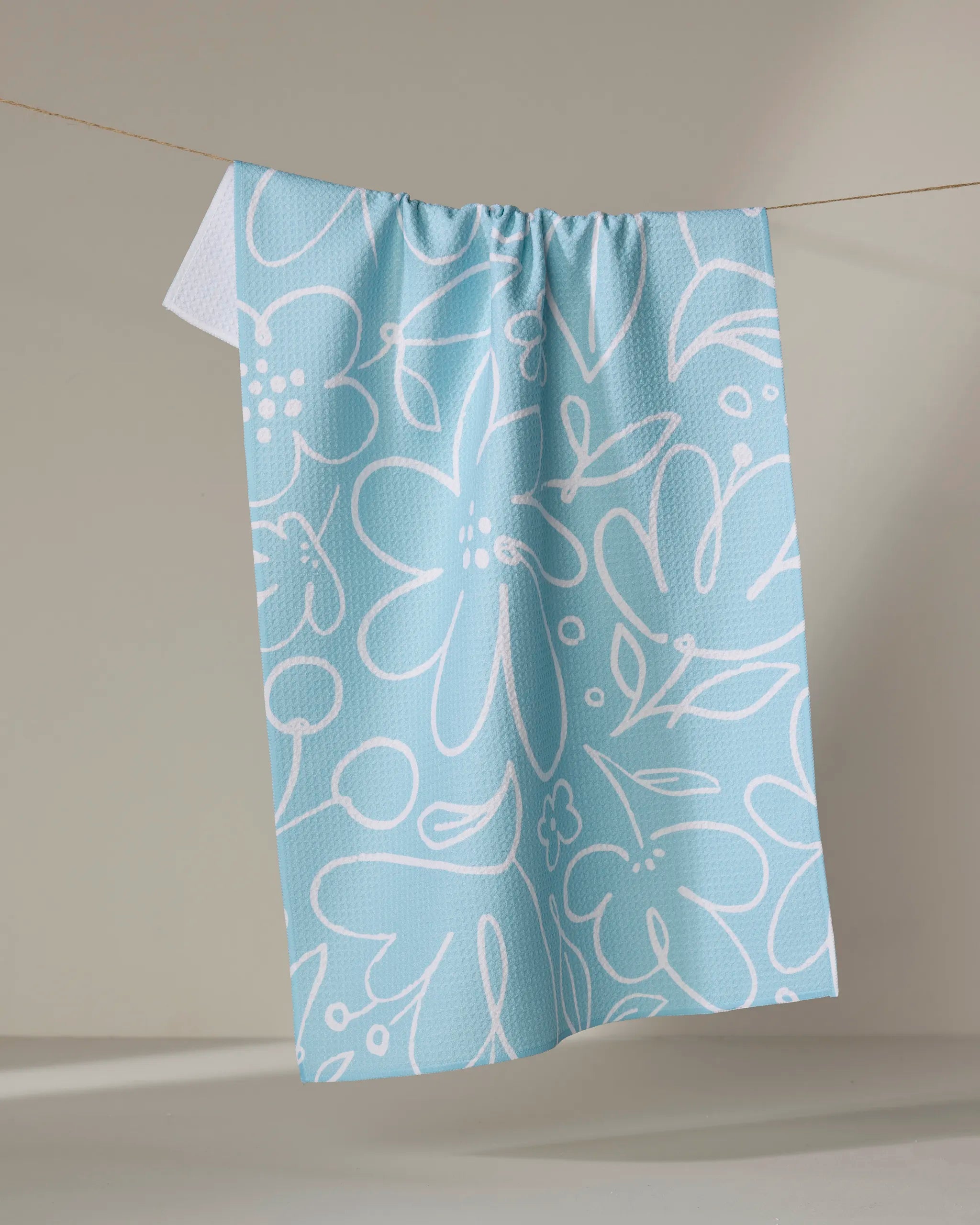 The Sara Mari "River Blossom" tea towel, featuring a white abstract floral pattern on light blue, hangs on a clothesline against a plain, neutral background.