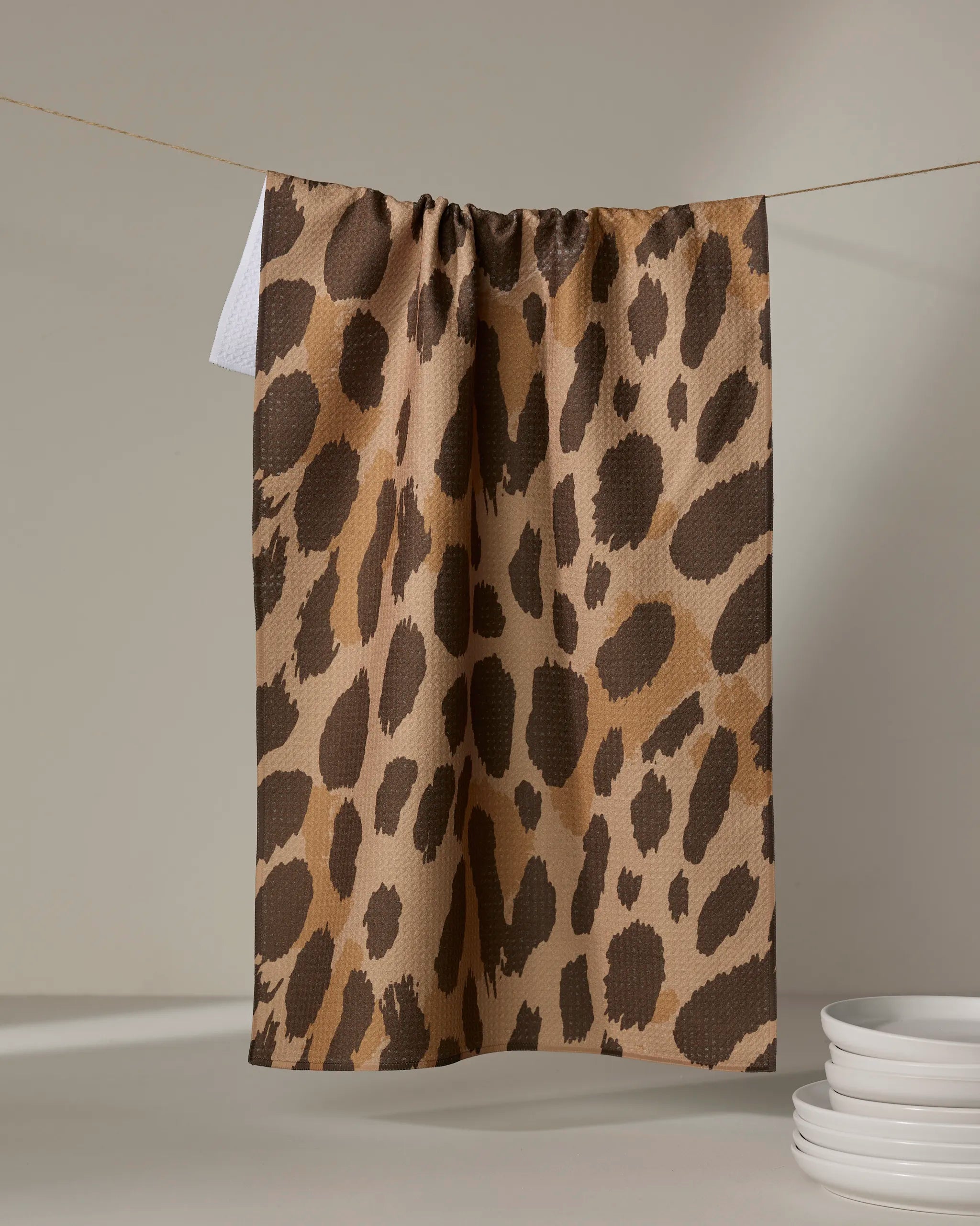 The Geometry Roam absorbent dish towel, featuring a brown and tan leopard print, hangs on a clothesline above a white surface with a small stack of white plates in the lower right corner.