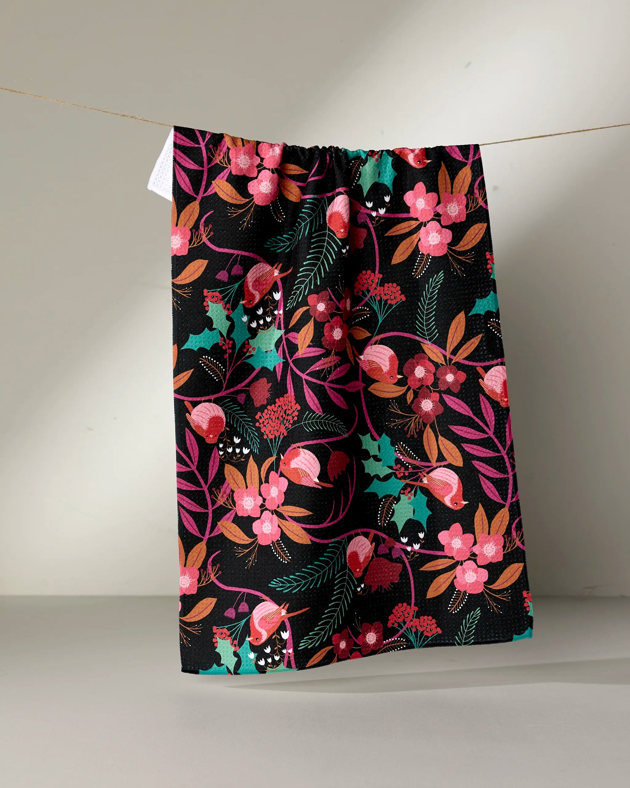A Rachel Parker "Robin Tree" tea towel, featuring a colorful floral and leaf pattern in pink, red, orange, teal, and green, hangs on a clothesline against a plain light background.