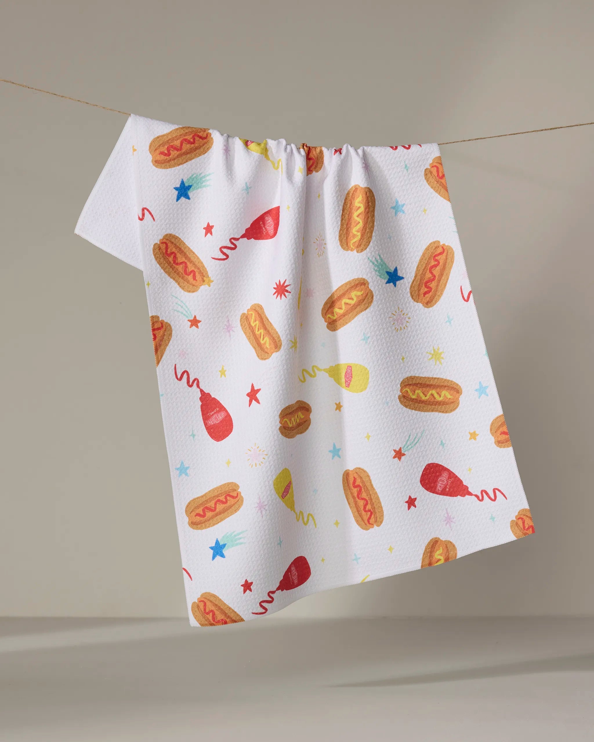 The "Rocket Relish" kitchen towel by Vaishali Design Studio hangs on a clothesline, featuring playful hot dogs, ketchup and mustard bottles, stars, and bursts on white recycled fabric—a festive touch for your kitchen.