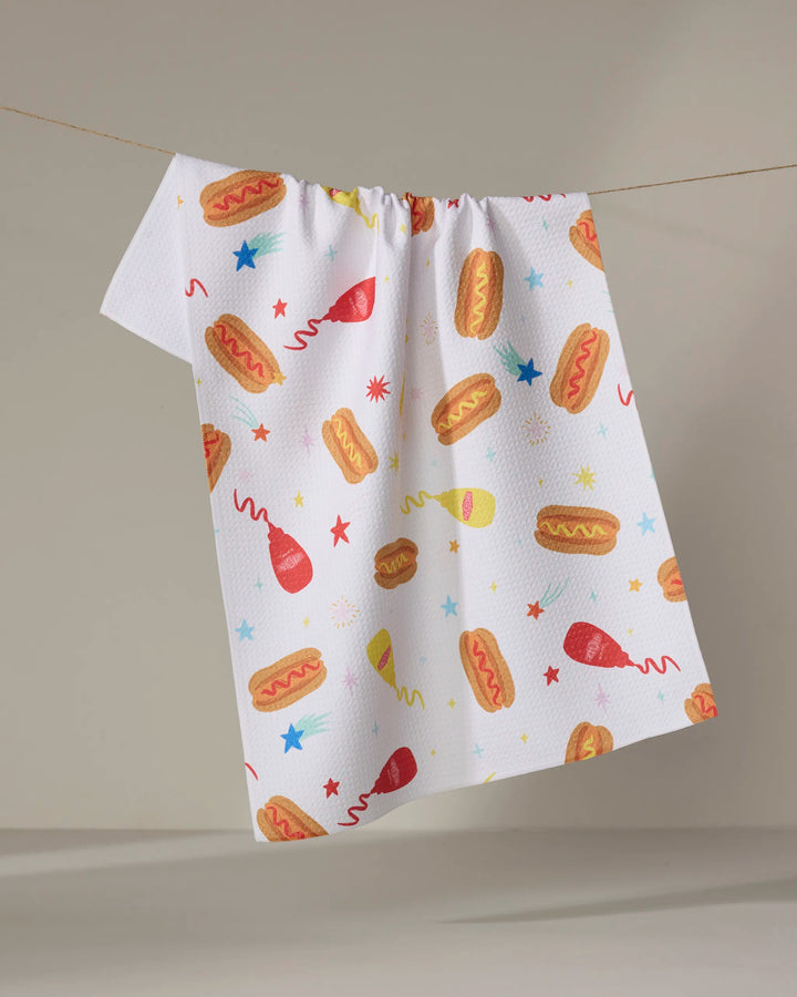 The "Rocket Relish" kitchen towel by Vaishali Design Studio hangs on a clothesline, featuring playful hot dogs, ketchup and mustard bottles, stars, and bursts on white recycled fabric—a festive touch for your kitchen.
