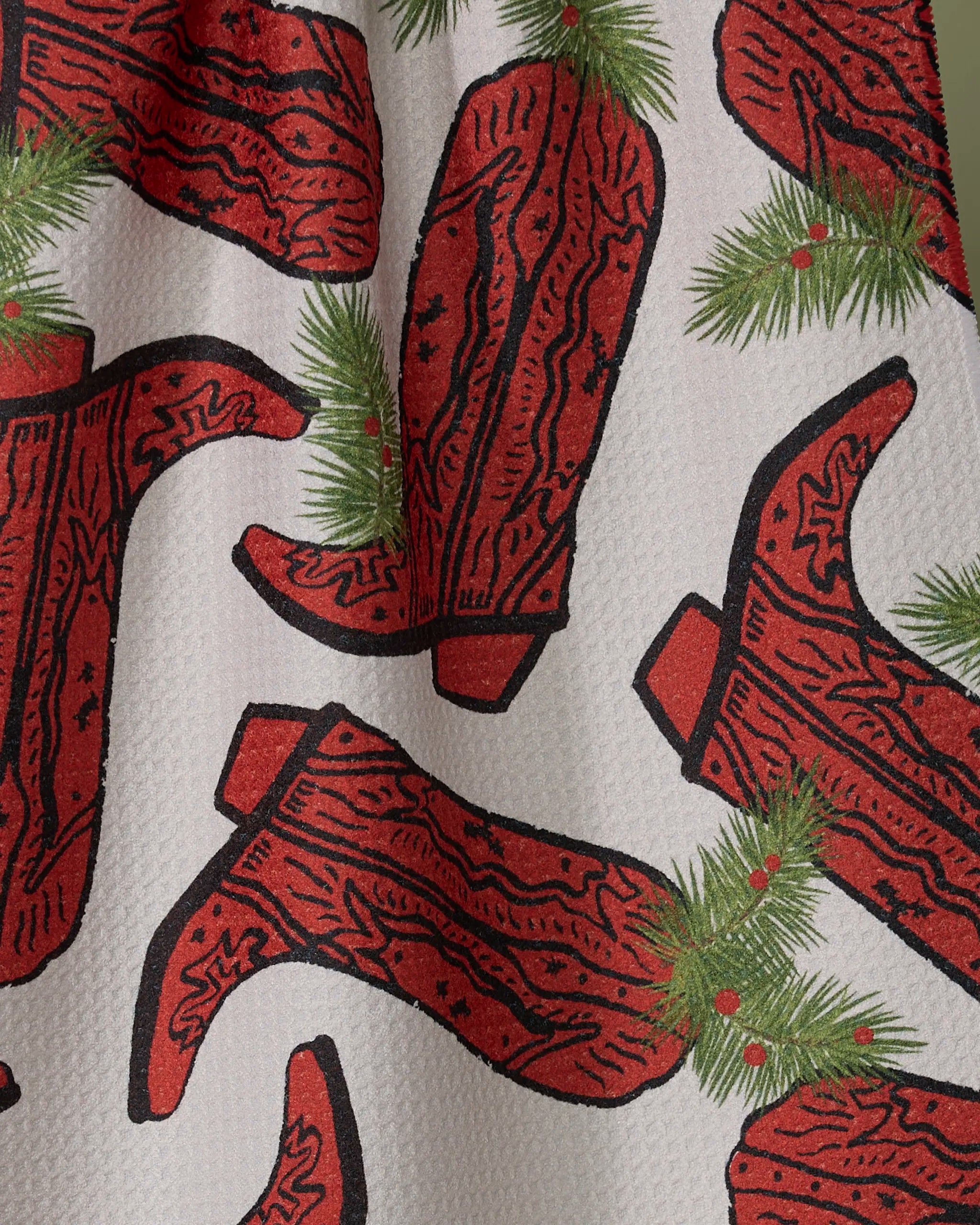 The Rodeo Red recycled kitchen towel by Geometry features red cowboy boots with black accents, green pine branches, and red berries on a textured white background.