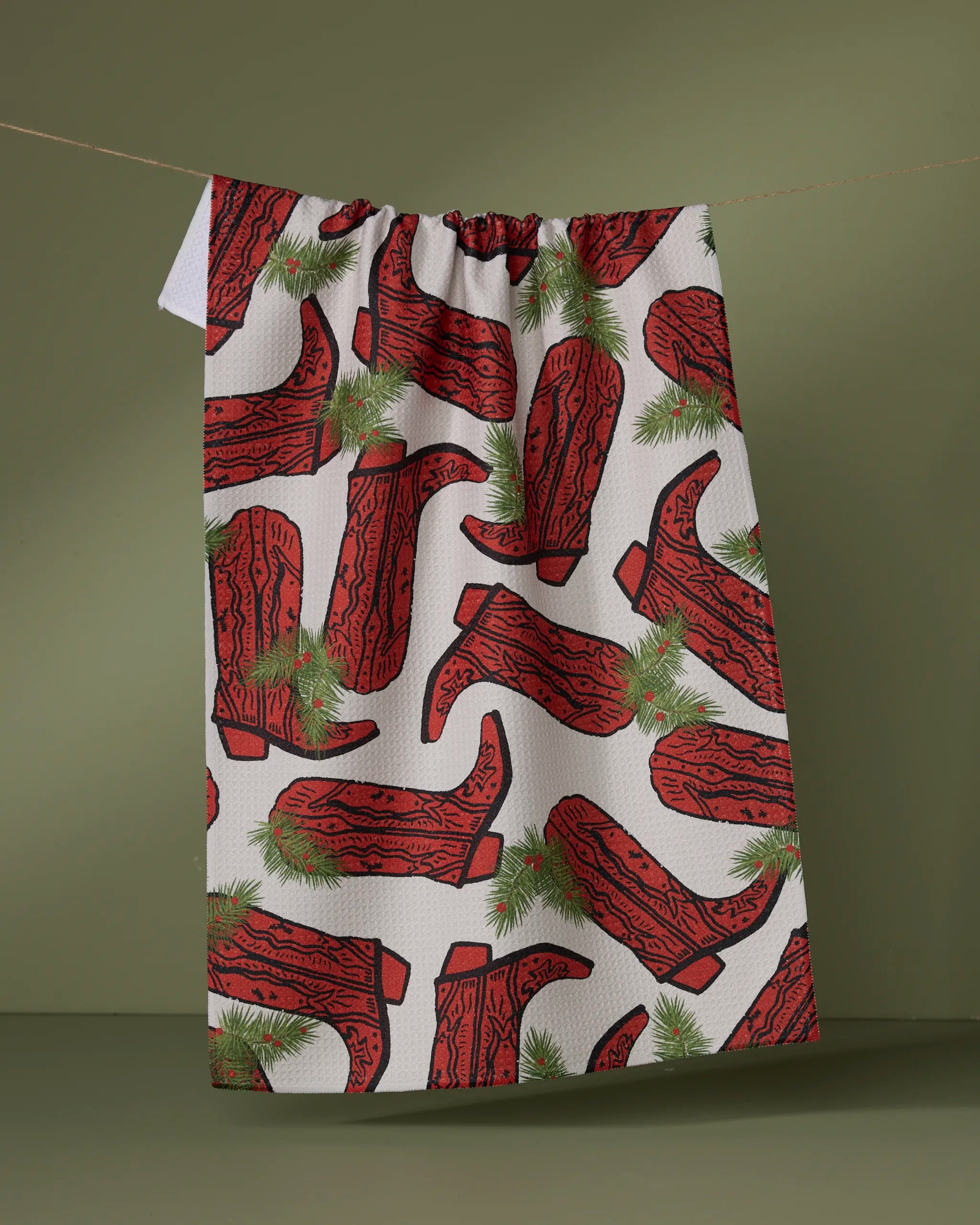 The Geometry Rodeo Red tea towel hangs on a string, displaying a pattern of red cowboy boots and green pine sprigs set against a light background.