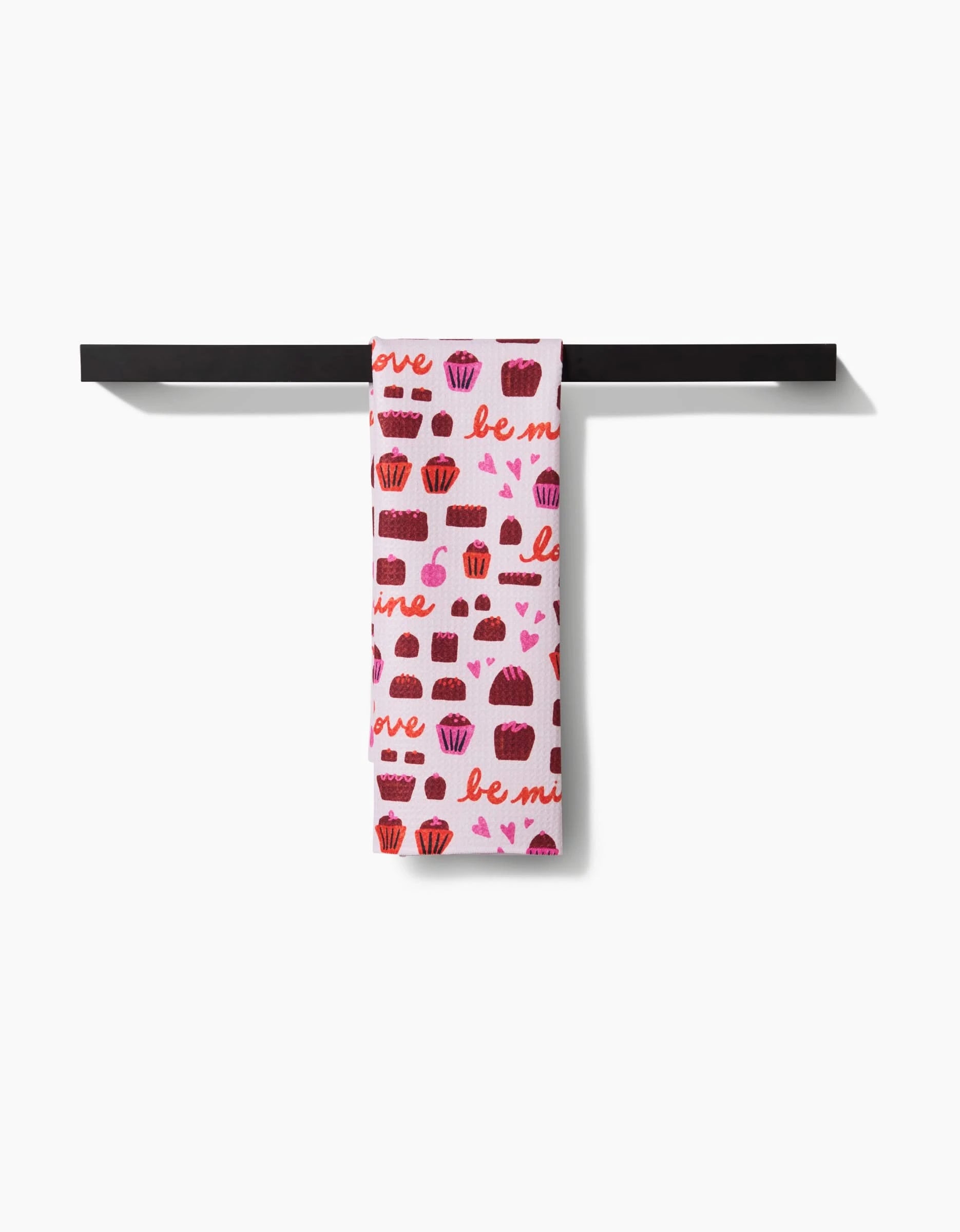The Amy MacCready Romeo white recycled kitchen towel, featuring red and pink cupcake, heart, and be mine patterns, hangs neatly on a black horizontal towel bar against a white background.