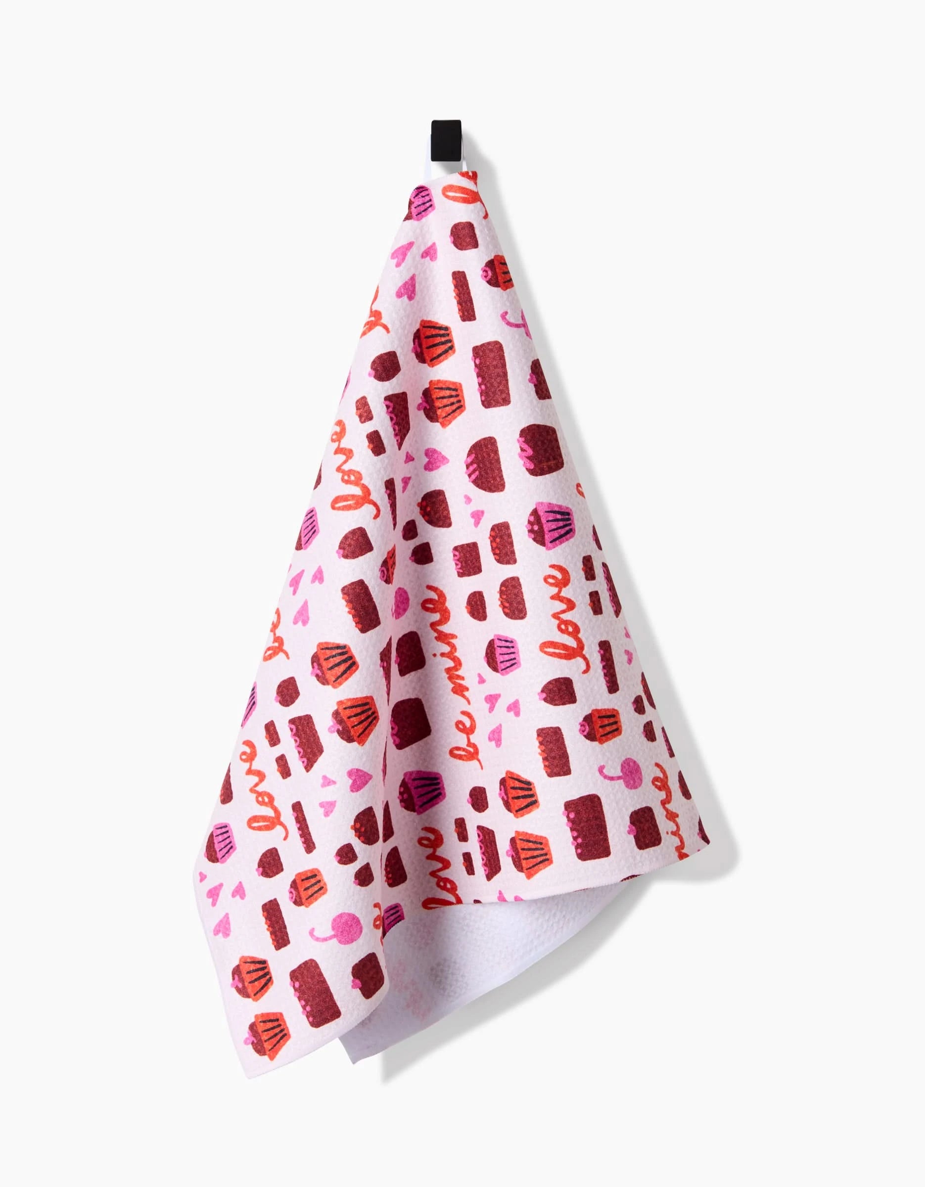 A white Romeo tea towel by Amy MacCready hangs on a black hook, featuring pink and red chocolates, hearts, and playful love script.
