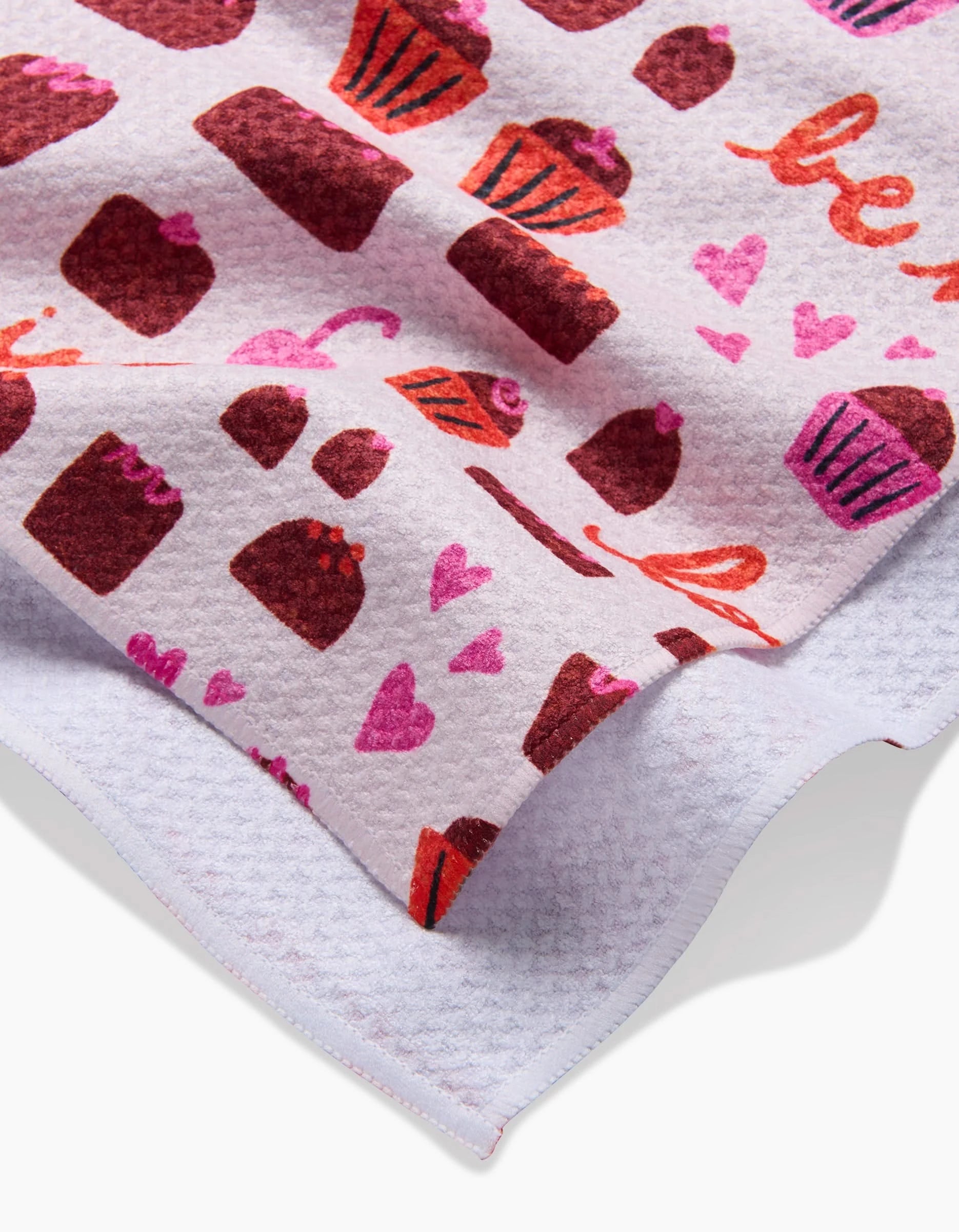 A close-up of the Amy MacCready Romeo quick-drying towel shows a pink background with red and pink cupcakes, hearts, and sweet treats. The towel displays a crisp white underside along the edge.