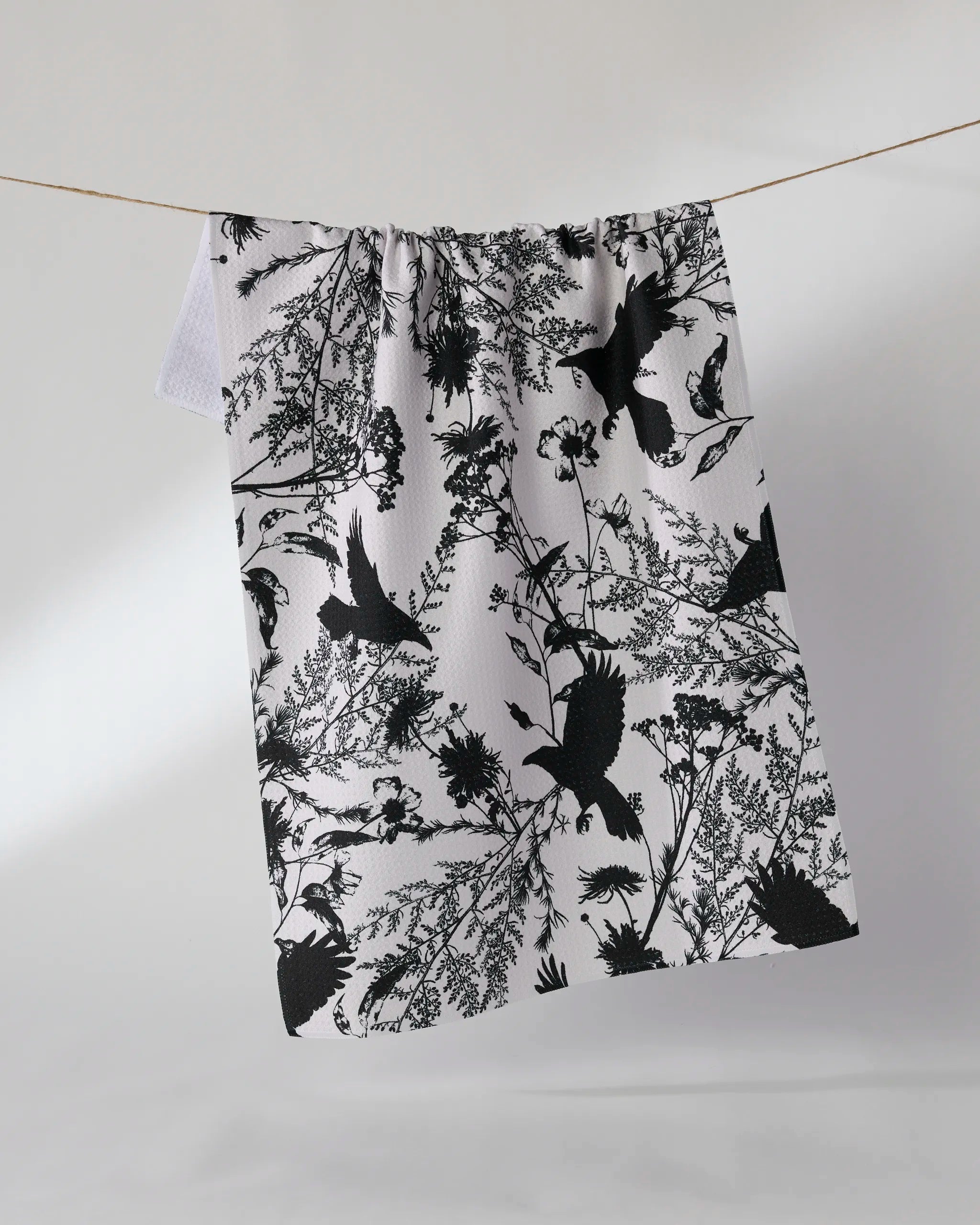 The Geometry Rookery kitchen towel, featuring a black botanical and bird silhouette design, hangs on a string against a plain light background.