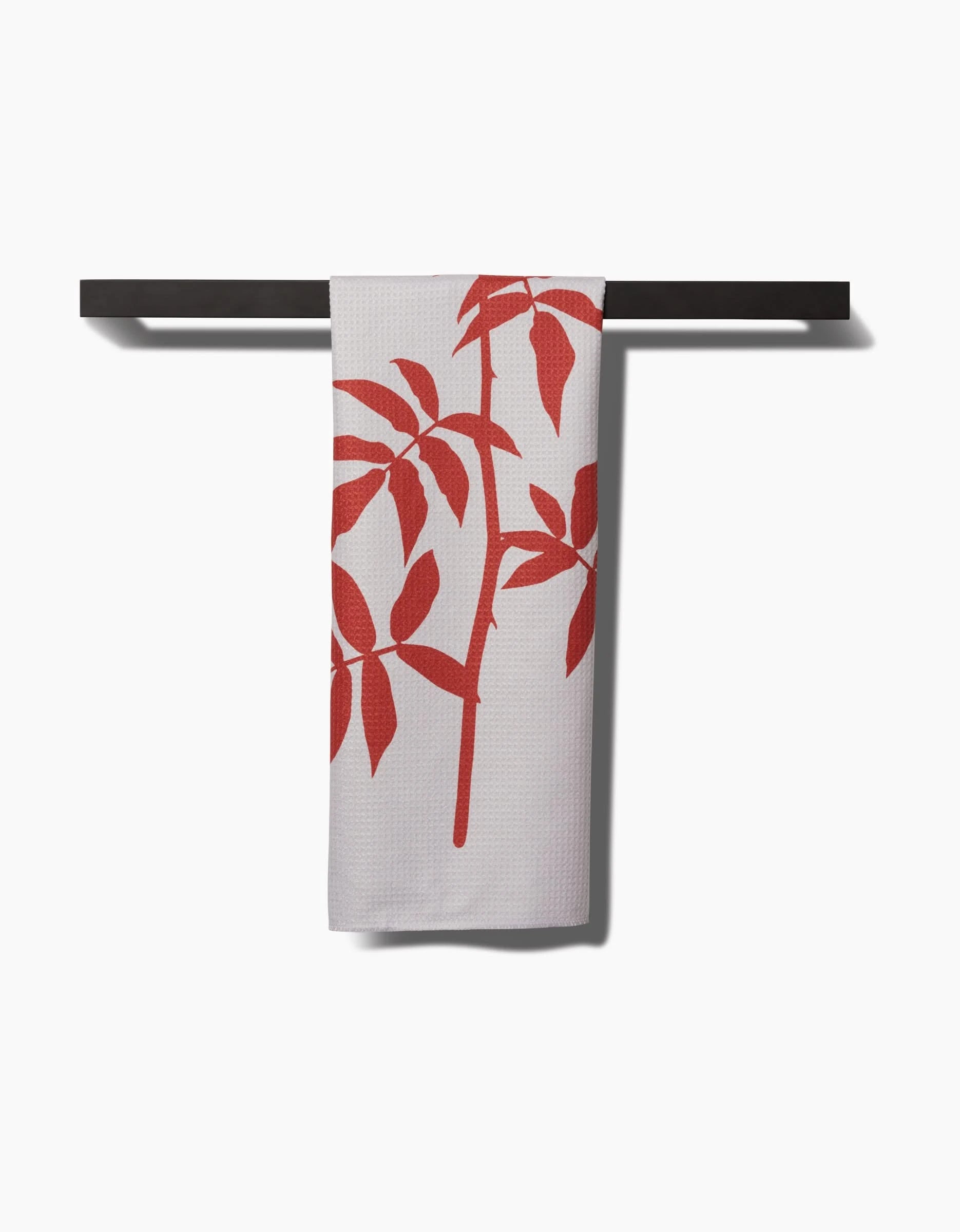 The Rose tea towel by PrintedPlant, made from recycled materials, displays a red leafy plant design and is draped over a black bar against a white background.