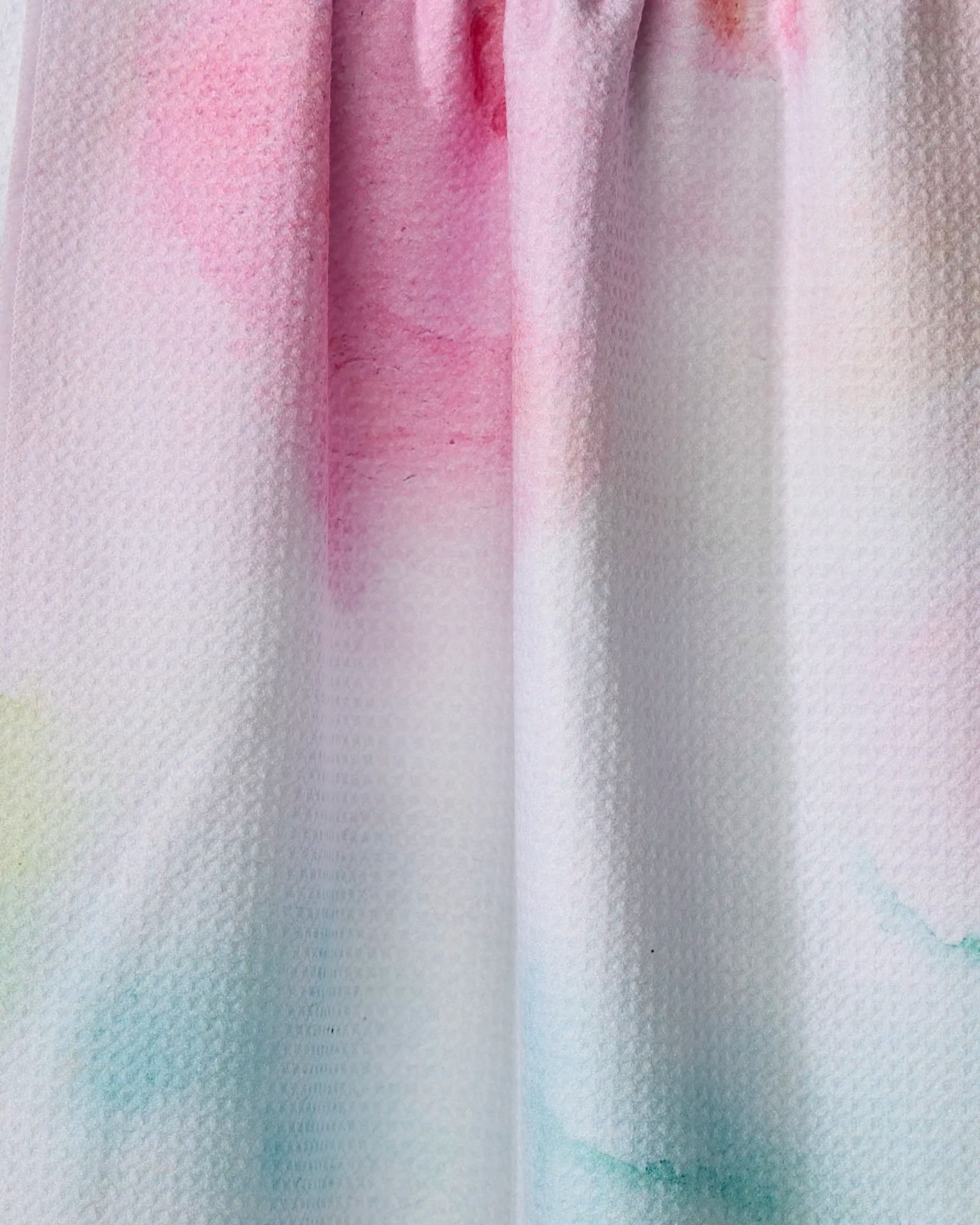 A close-up of the Stefanie Bales Rose Garden tea towel, made from recycled materials, showcases soft pastel watercolor patterns in pink, green, and blue with gentle folds and a subtle honeycomb texture.