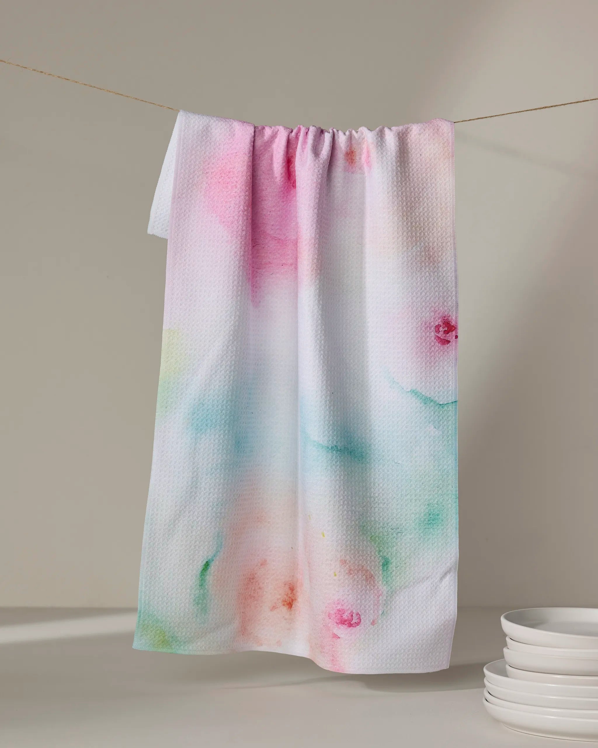 The Rose Garden tea towel by Stefanie Bales, featuring a pastel watercolor design and made from recycled materials, hangs on a clothesline above white plates against a neutral backdrop.