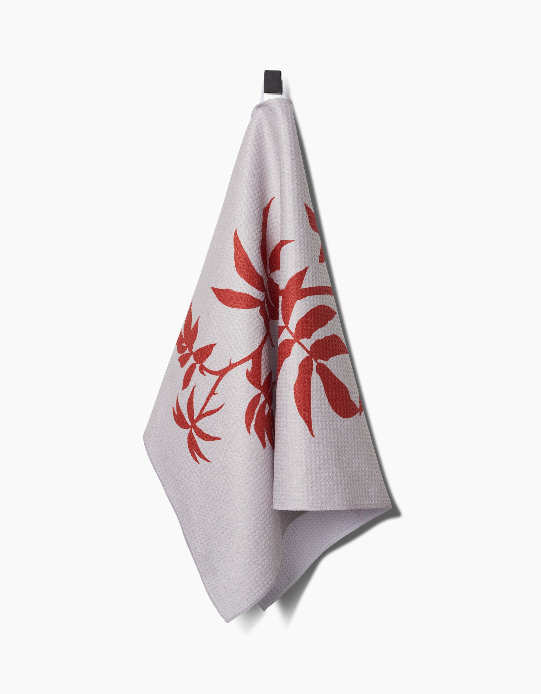The PrintedPlant Rose tea towel, featuring a red floral pattern on white textured fabric made from recycled materials, hangs on a black hook against a plain white background.