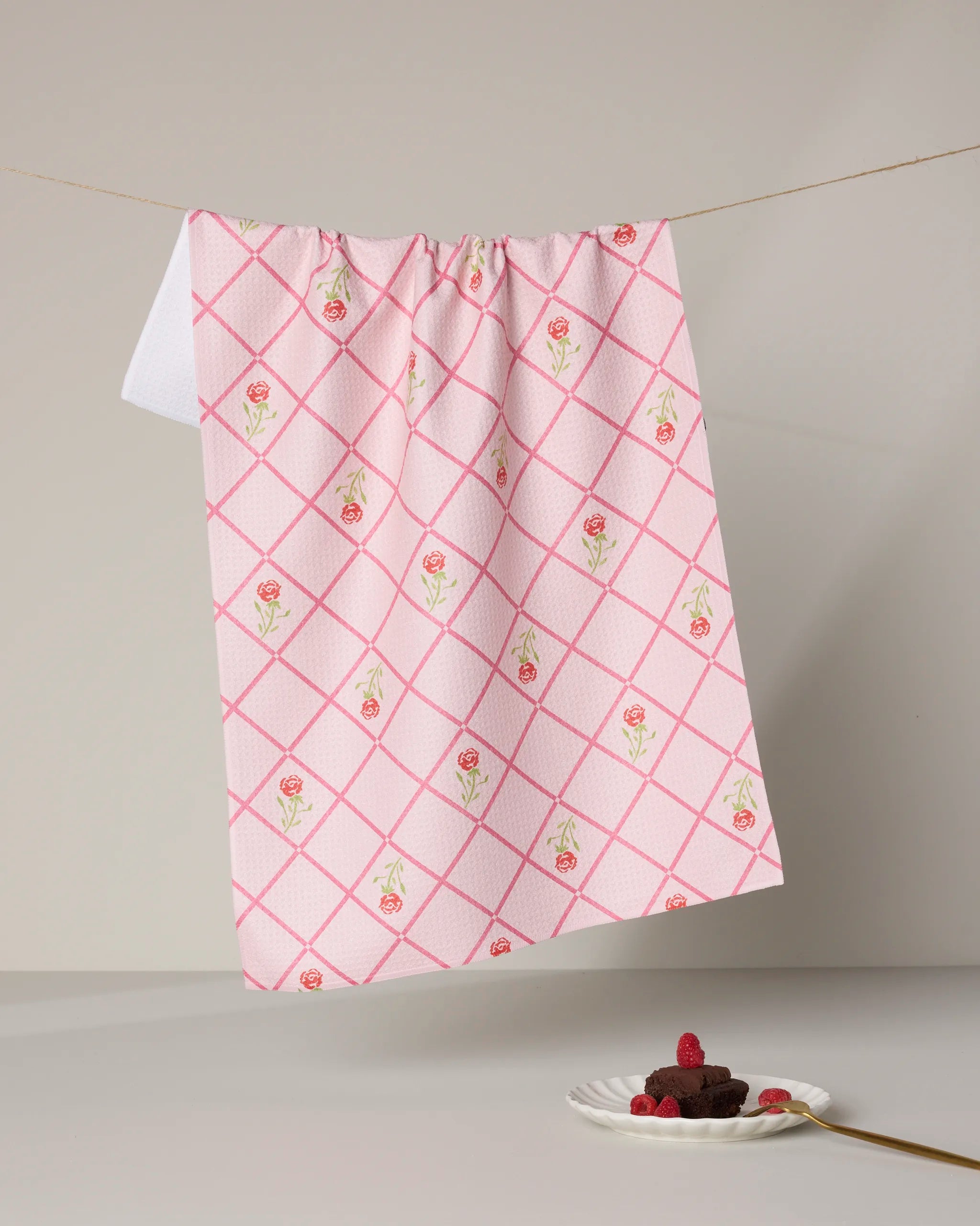The Geometry Rose Lattice tea towel, featuring a pink and white rose-checkered design, hangs on a clothesline above a plate of brownies with raspberries and a gold spoon on a neutral backdrop.