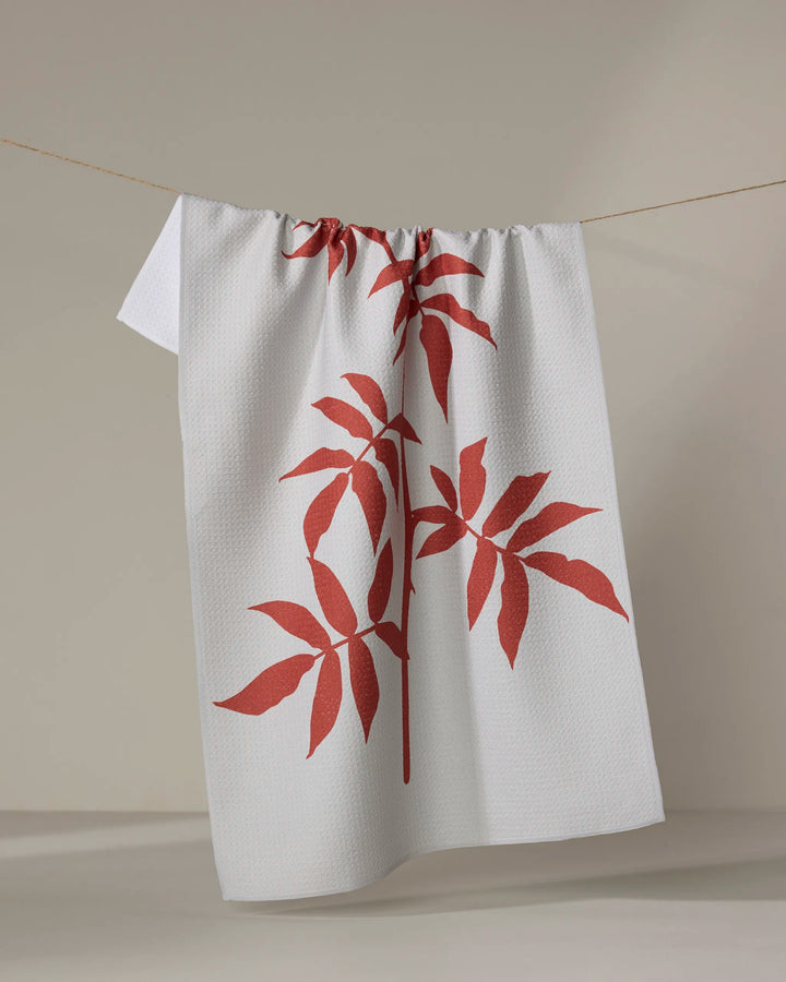 The "Rose" tea towel by PrintedPlant, featuring two large red leafy plant designs, hangs on a clothesline against a plain, light background.