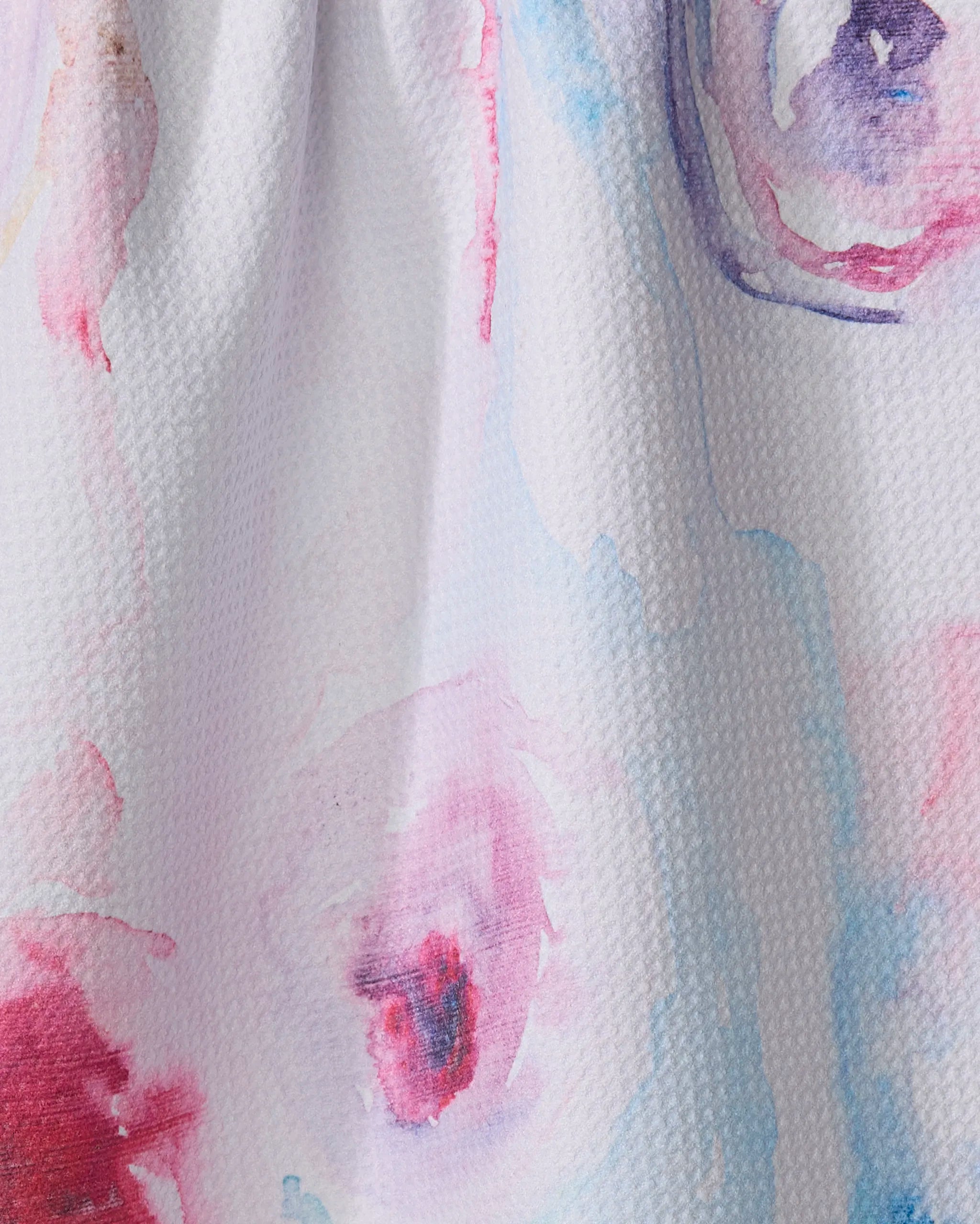 A close-up of Stefanie Bales' "Roses" kitchen towel shows its textured white recycled fabric with soft watercolor-style pink, purple, and blue splotches blending into an artistic abstract pattern.