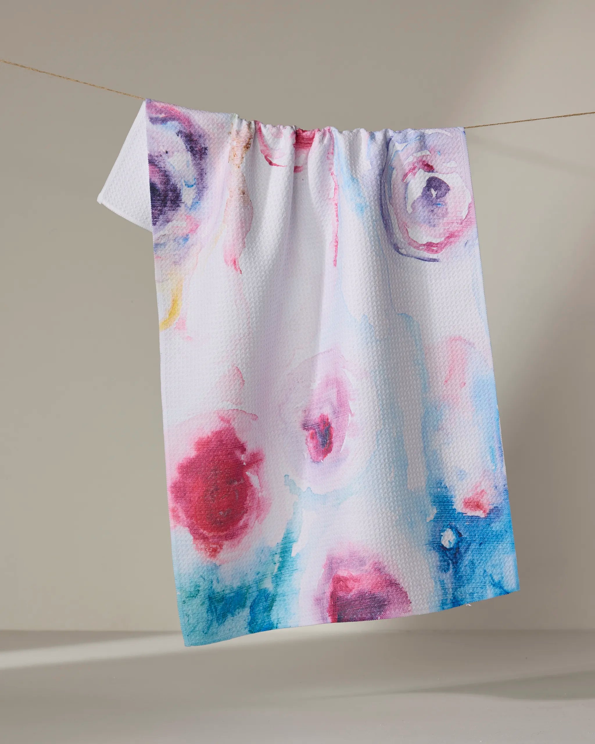 The Roses towel by Stefanie Bales is ultra-absorbent, white, and features colorful abstract watercolor-like spots in red, blue, and purple. It hangs on a clothesline against a neutral background.