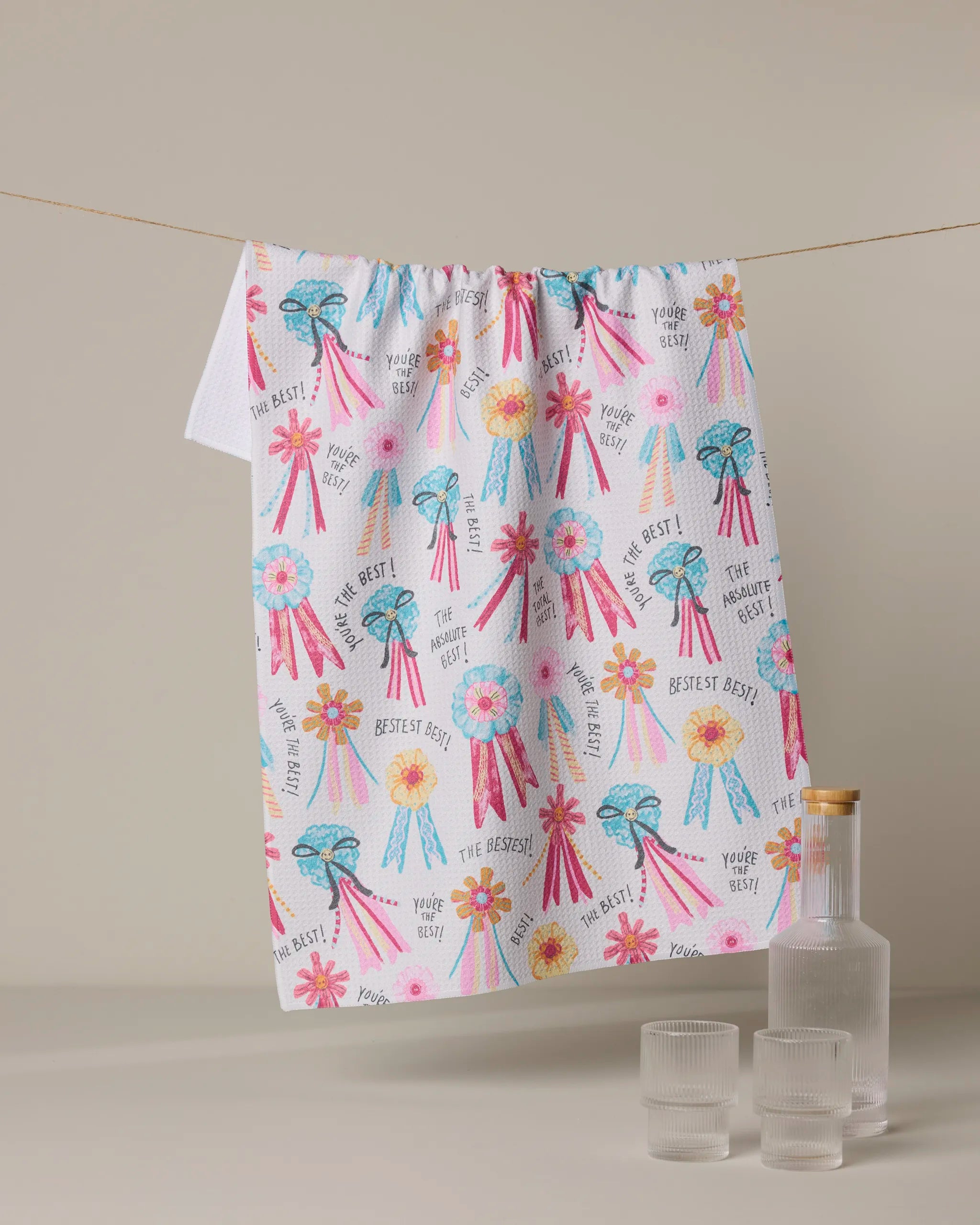 The Geometry Rosettes for You tea towel, made from recycled materials and decorated with award ribbons, flowers, and phrases like "You’re the best!" and "The greatest!", hangs above a neutral surface with two glass cups and a carafe.