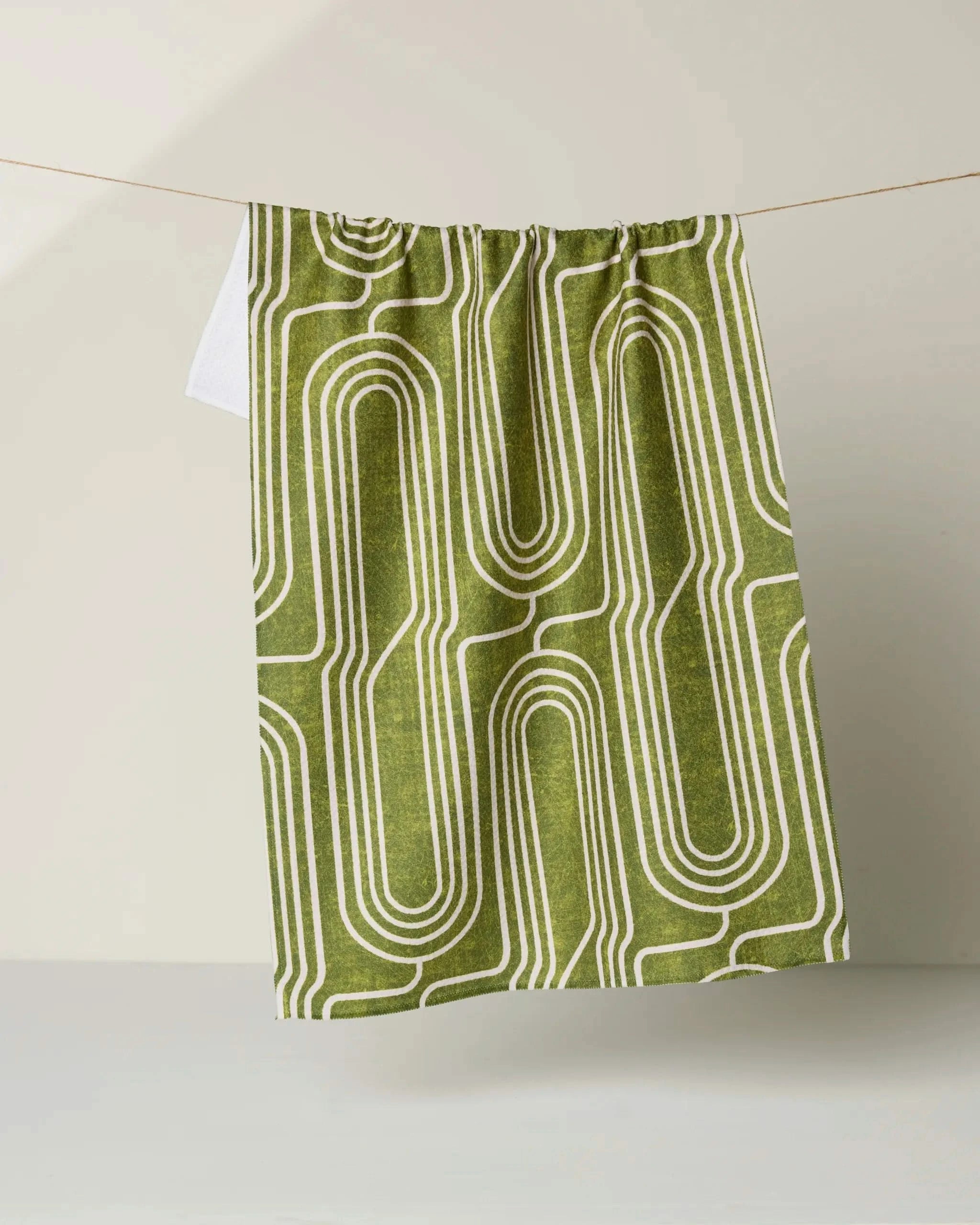 The Roslyn green tea towel by Amy MacCready is made from recycled materials and features a white geometric maze pattern, shown hanging on a clothesline against a light background.
