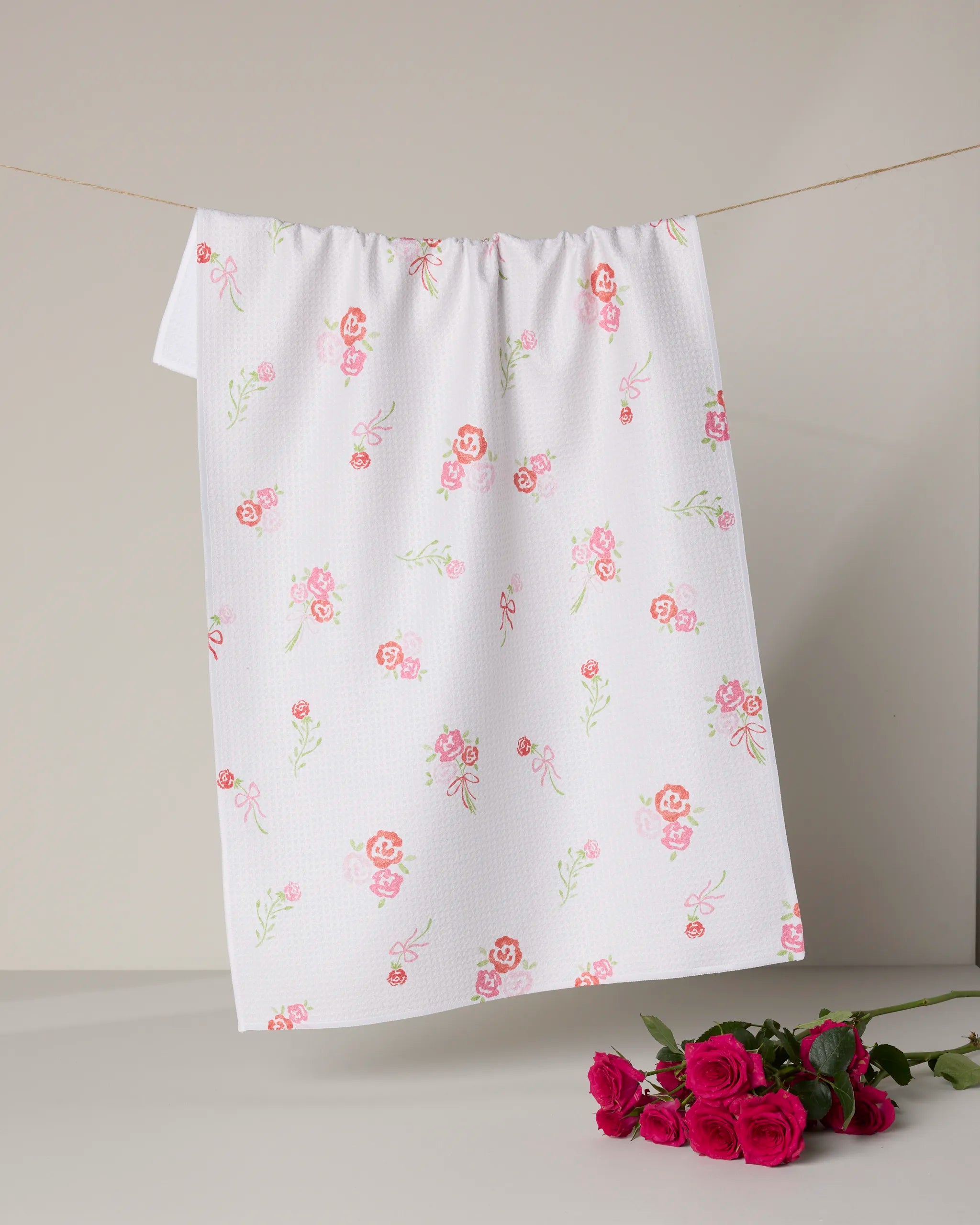 The Rosy Cheeks tea towel by Geometry, made from recycled materials, features a pink and green floral pattern. Hung against a light background, it’s both charming and eco-friendly. A small bouquet of pink roses sits below the towel.
