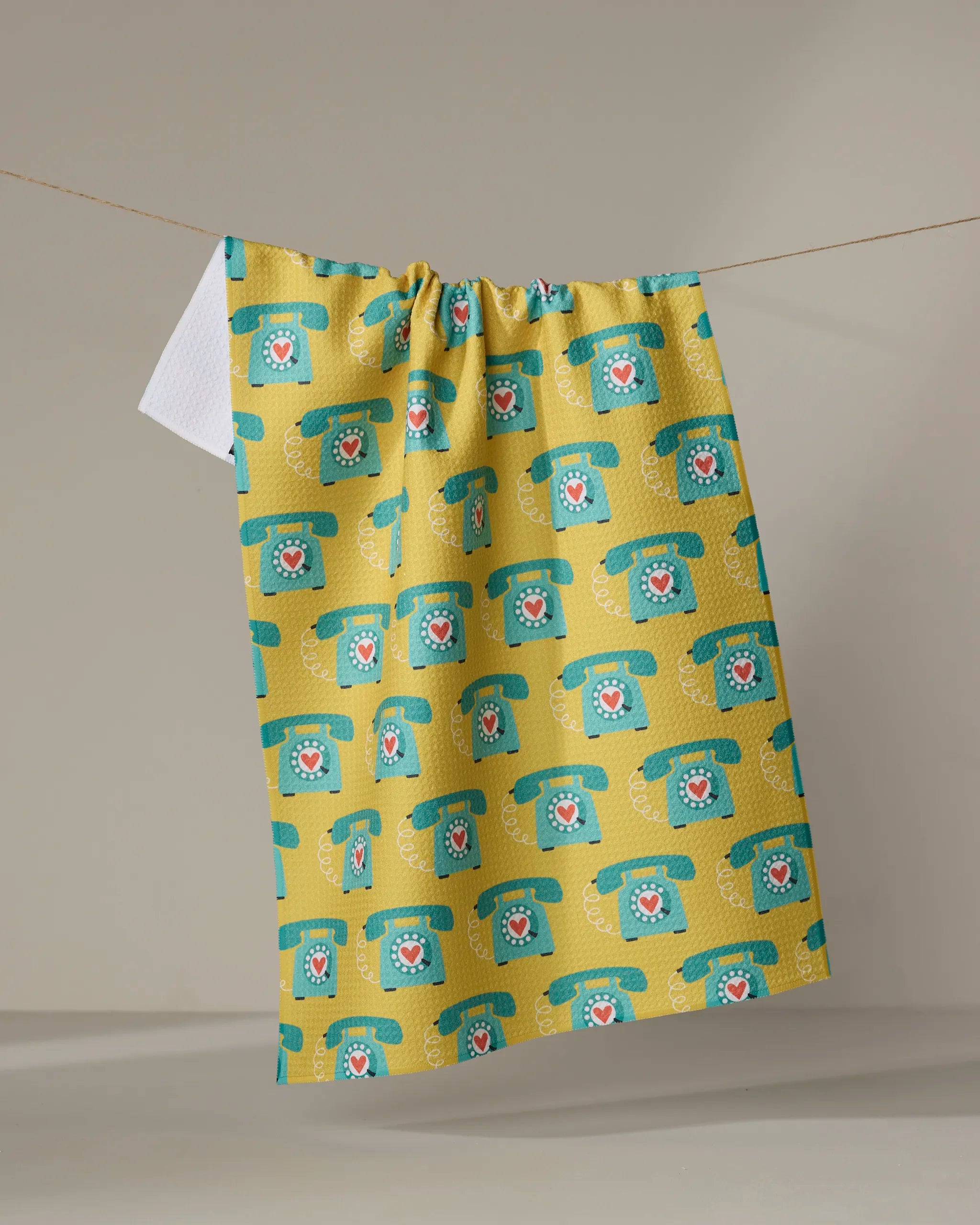 The "Rotary Phones" kitchen towel by Megan Kampa features a yellow background with vintage turquoise rotary phones and heart centers—an absorbent, charming tea towel for any home, shown hanging on a beige background.