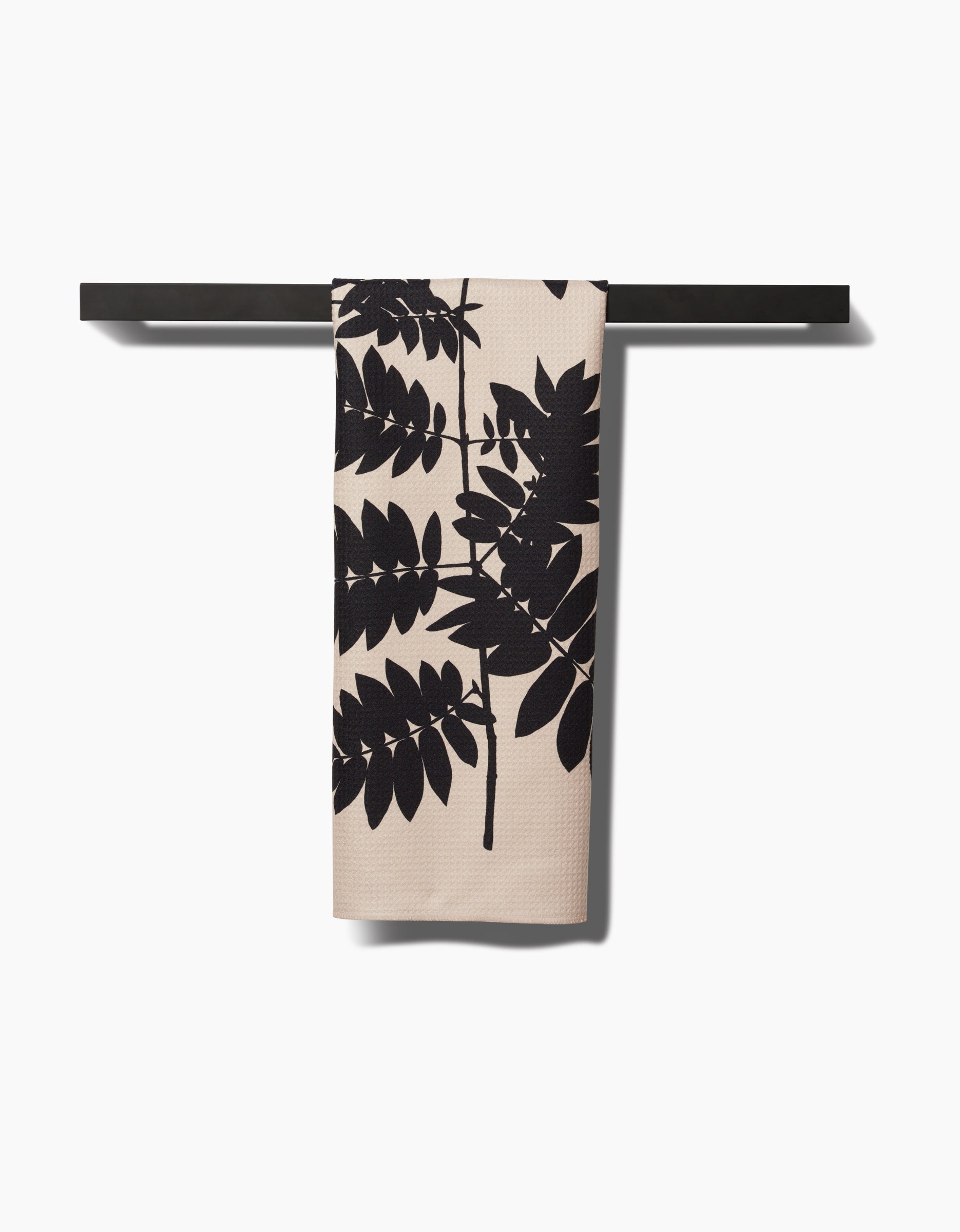 The Rowan tea towel by PrintedPlant features a bold black leafy branch pattern on beige, crafted from ultra-absorbent Geoweave fabric, and hangs neatly over a black bar against a white background.