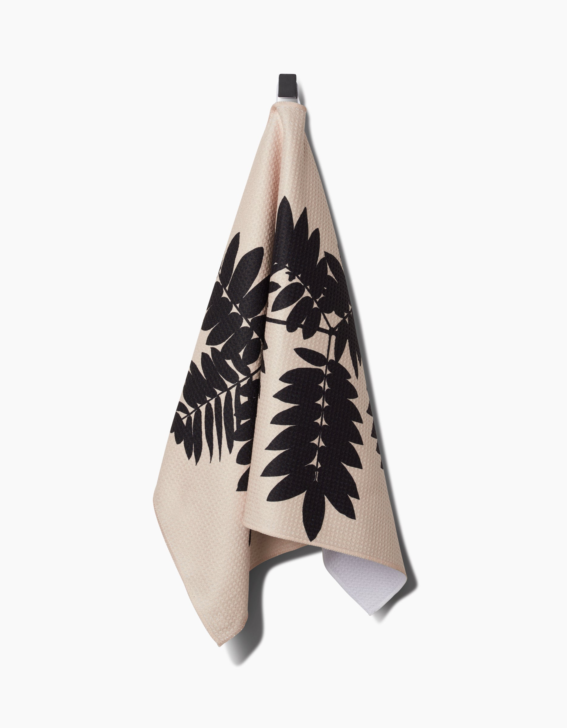 The Rowan tea towel by PrintedPlant features a large black leaf pattern on beige, hanging from a hook against a white background. Its made from ultra-absorbent Geoweave fabric.