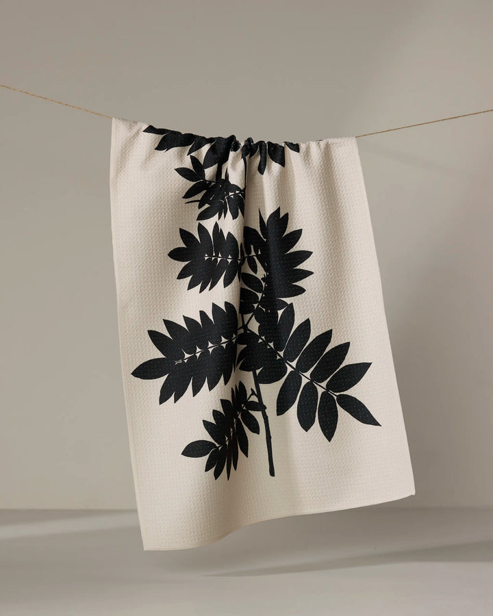The Rowan tea towel by PrintedPlant, made from recycled materials, features a bold black botanical leaf print on beige and is shown hanging on a clothesline against a plain light background.