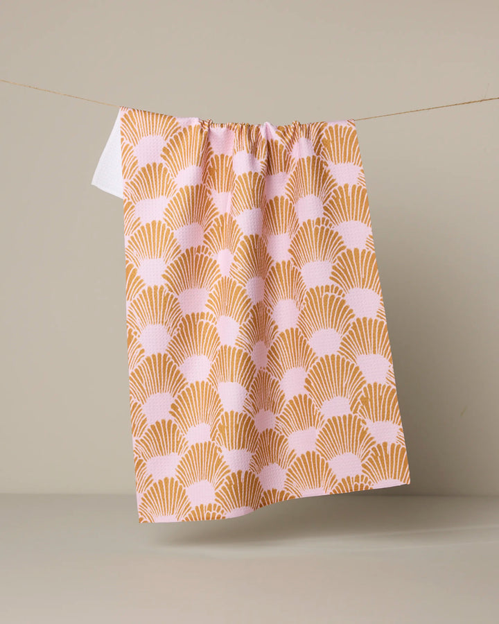 The Geometry Royal Scallop kitchen tea towel, featuring a repeating pink and mustard yellow fan pattern and made from recycled materials, hangs against a plain light background.
