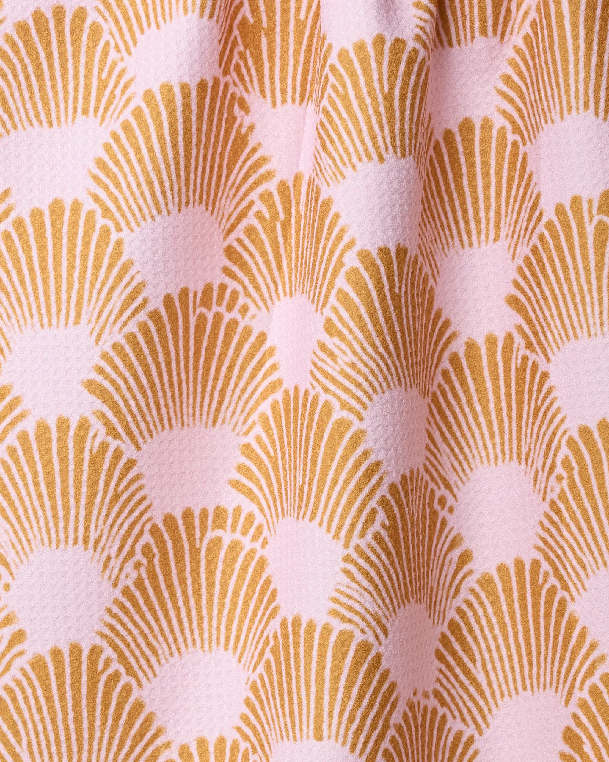 A close-up of the Geometry Royal Scallop tea towel shows its absorbent, slightly raised texture and wavy feel, featuring a repeating gold fan-shaped pattern on a light pink background.
