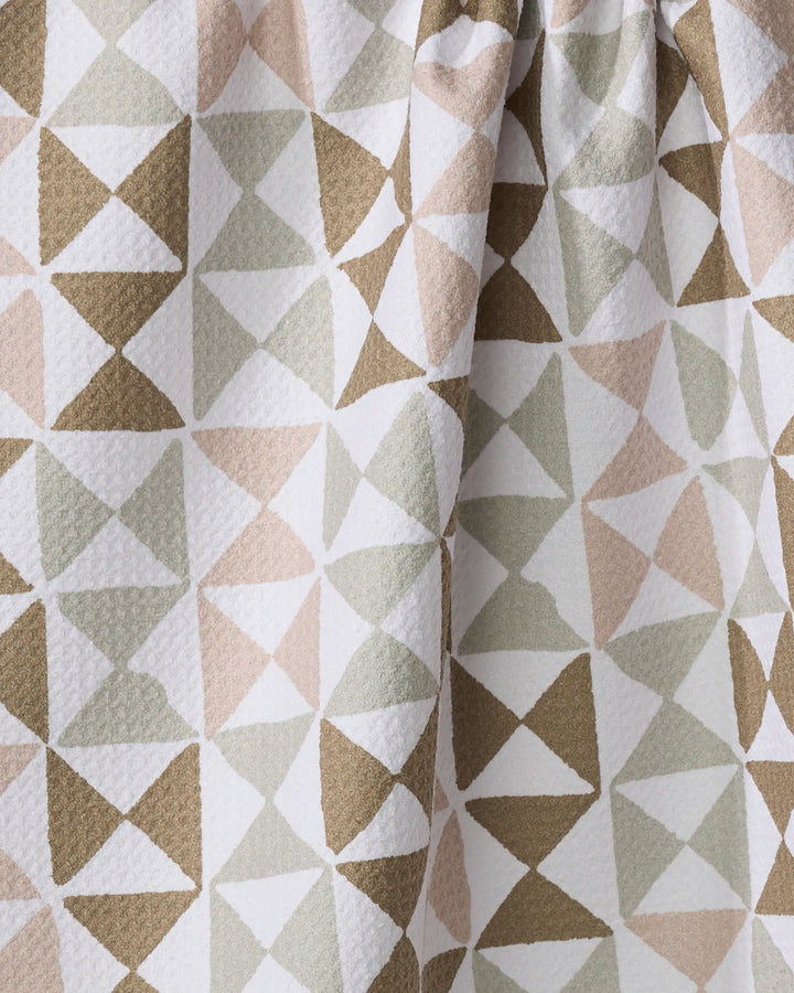 Close-up of the Rubix tea towel by Studio Jeannot, featuring a geometric pattern of overlapping triangles in beige, taupe, olive green, and white. The textured fabric is slightly gathered at the top.