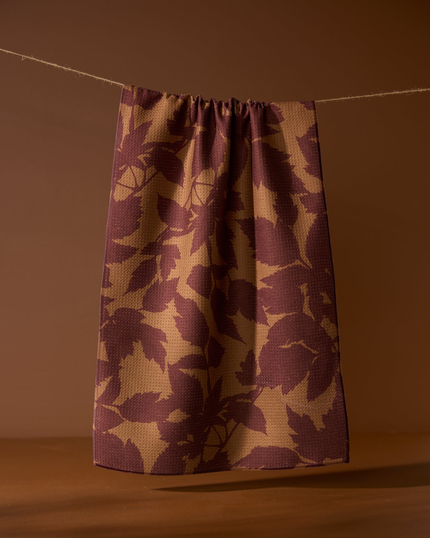 The Rustle tea towel by Geometry, featuring a dark red leaf pattern on brown and made from recycled materials, hangs on a string against a brown background.