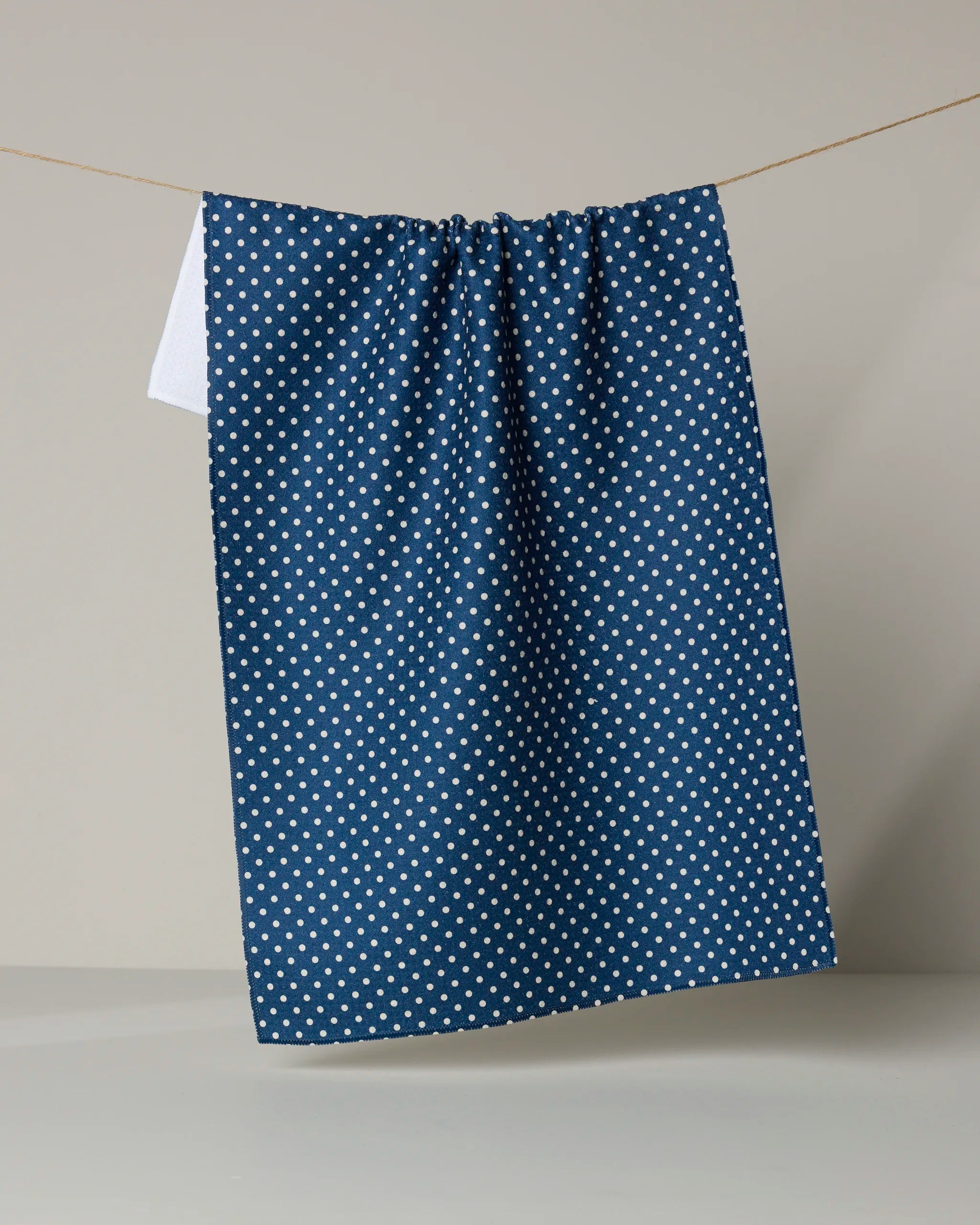 The Saddle Indigo tea towel by Geometry, featuring quick-drying recycled material and white polka dots, hangs on a clothesline against a beige backdrop; a hint of white cloth peeks out from behind the blue fabric.