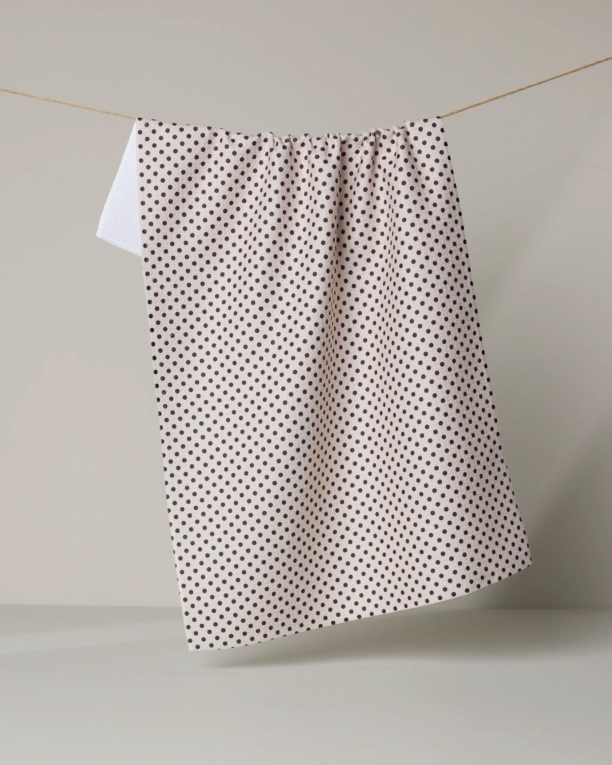 The Geometry Saddle Off White recycled kitchen towel with small black polka dots hangs over a clothesline against a plain, light gray background.