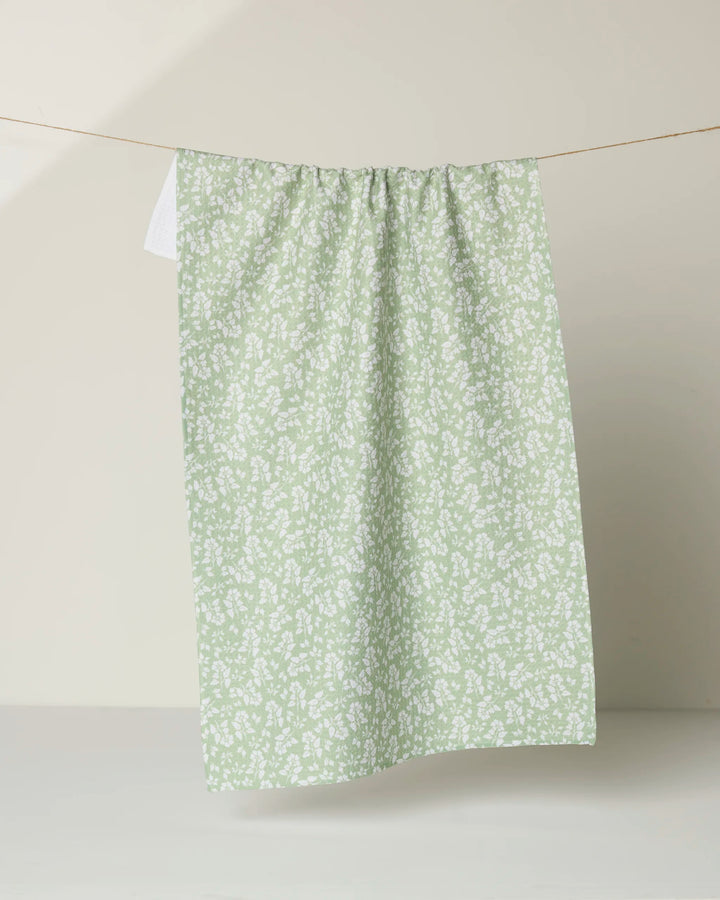 The Danelys Sidron "Sage Garden" recycled kitchen towel, featuring a white floral pattern, hangs on a string against a neutral beige background.