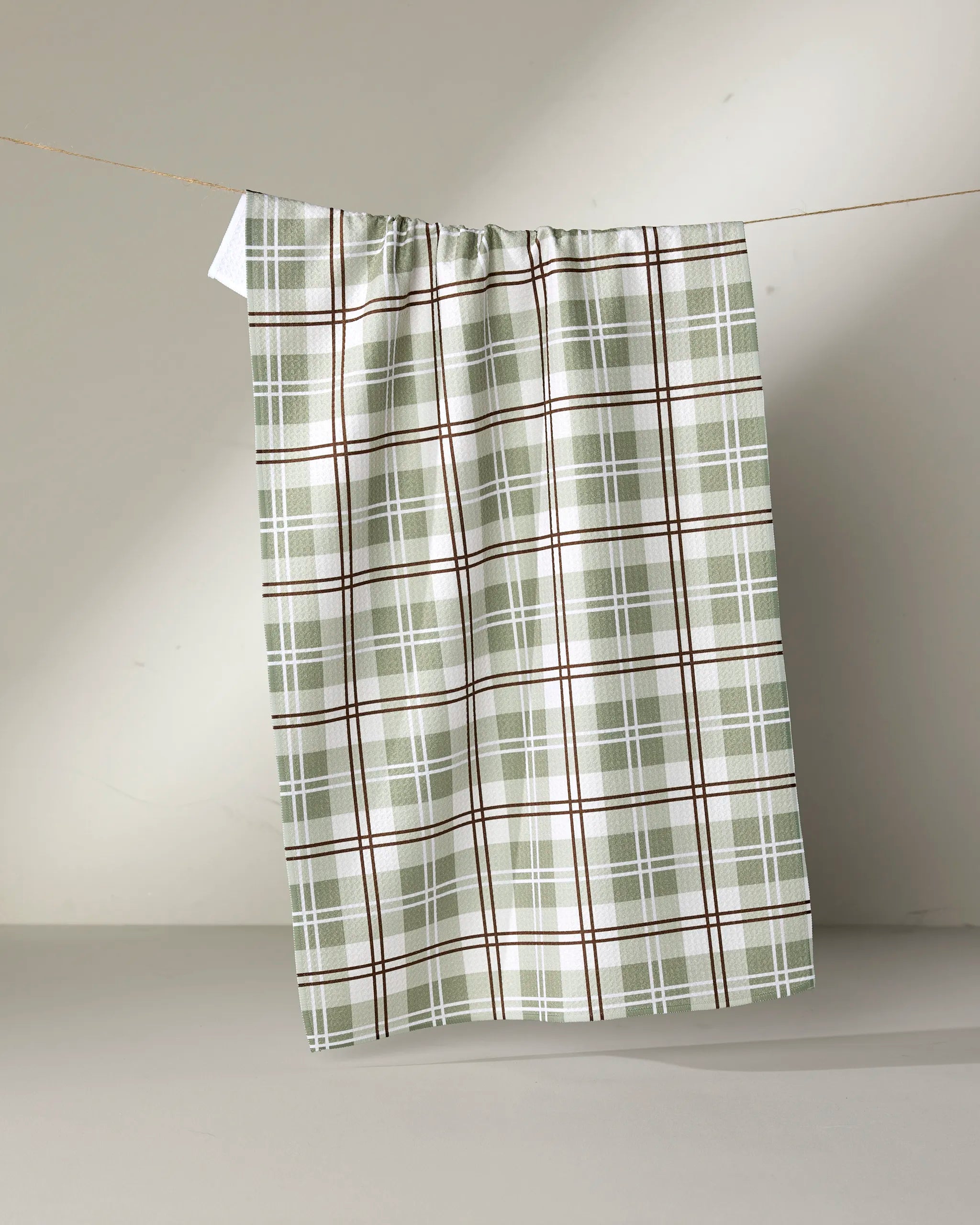 The Sage Season tea towel by Ashley Reeves, crafted from recycled materials in a green, white, and brown plaid pattern, hangs on a thin string against a neutral gray background.