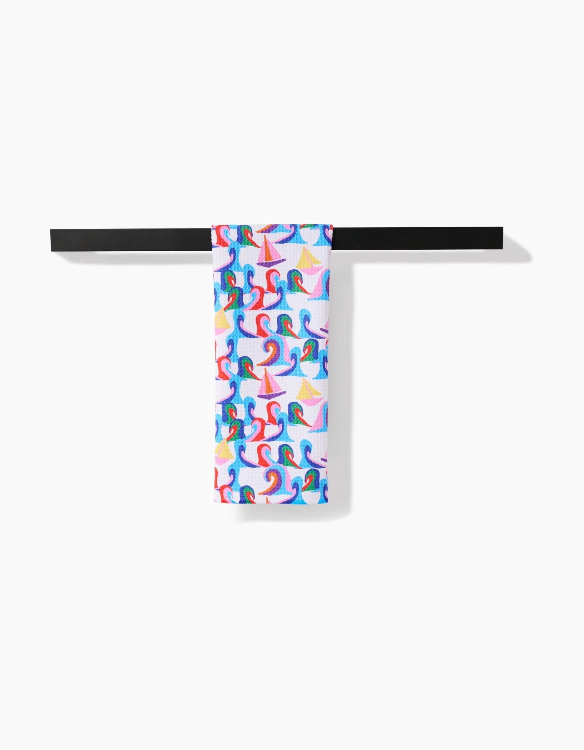 The Sail Safe towel by Ania Zwara Art, featuring a vibrant pattern of stylized surfers on waves and made from ultra-absorbent Geoweave fabric, hangs neatly on a black horizontal towel bar against a white background.