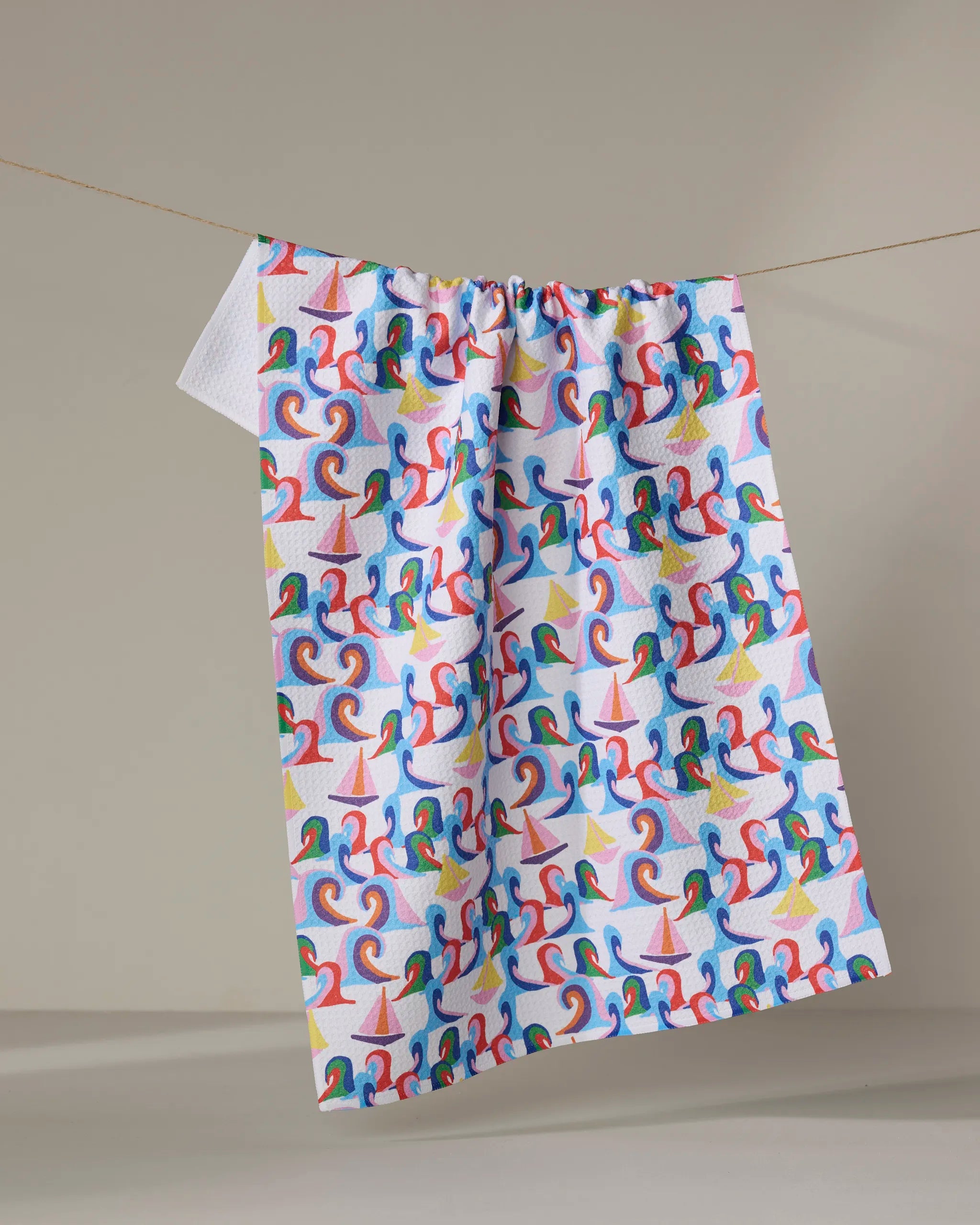 The Sail Safe quick-drying towel by Ania Zwara Art features an abstract sailboat pattern in red, blue, yellow, and green and hangs on a clothesline against a neutral background.