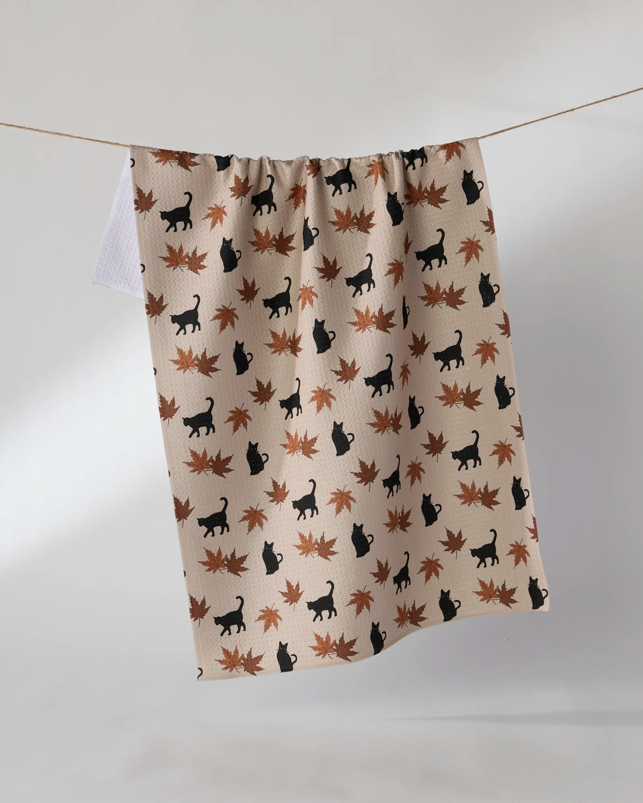 The Geometry "Salem Woods" tea towel, made from recycled materials, features black cat silhouettes and orange-brown maple leaves on beige fabric and hangs on a string against a plain white background.