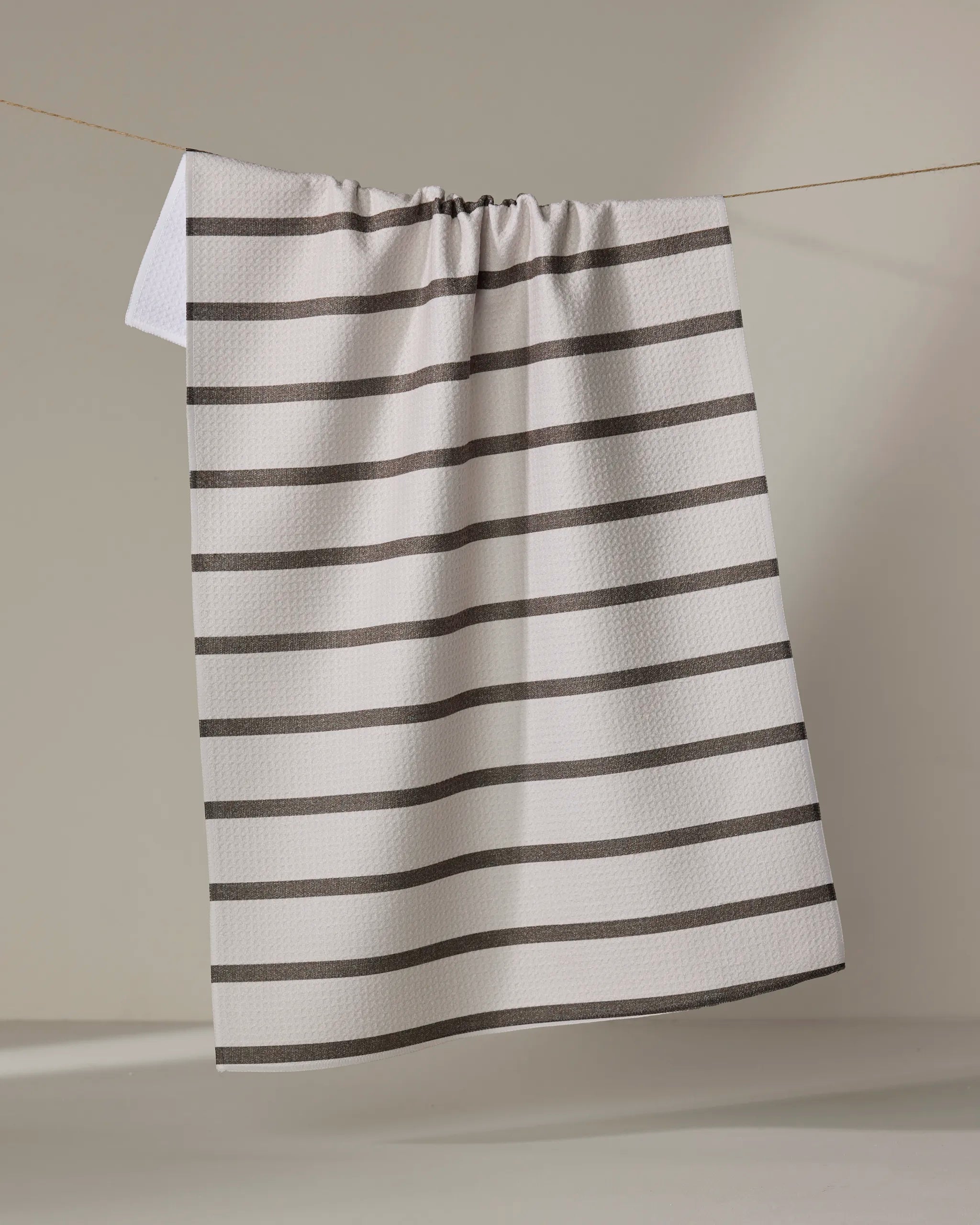 The Jenny Keller Salt Air Stripe tea towel, featuring quick-drying white fabric with evenly spaced horizontal black stripes, hangs on a clothesline against a neutral background.