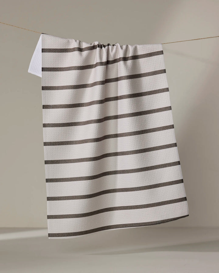 The Jenny Keller Salt Air Stripe tea towel, featuring quick-drying white fabric with evenly spaced horizontal black stripes, hangs on a clothesline against a neutral background.