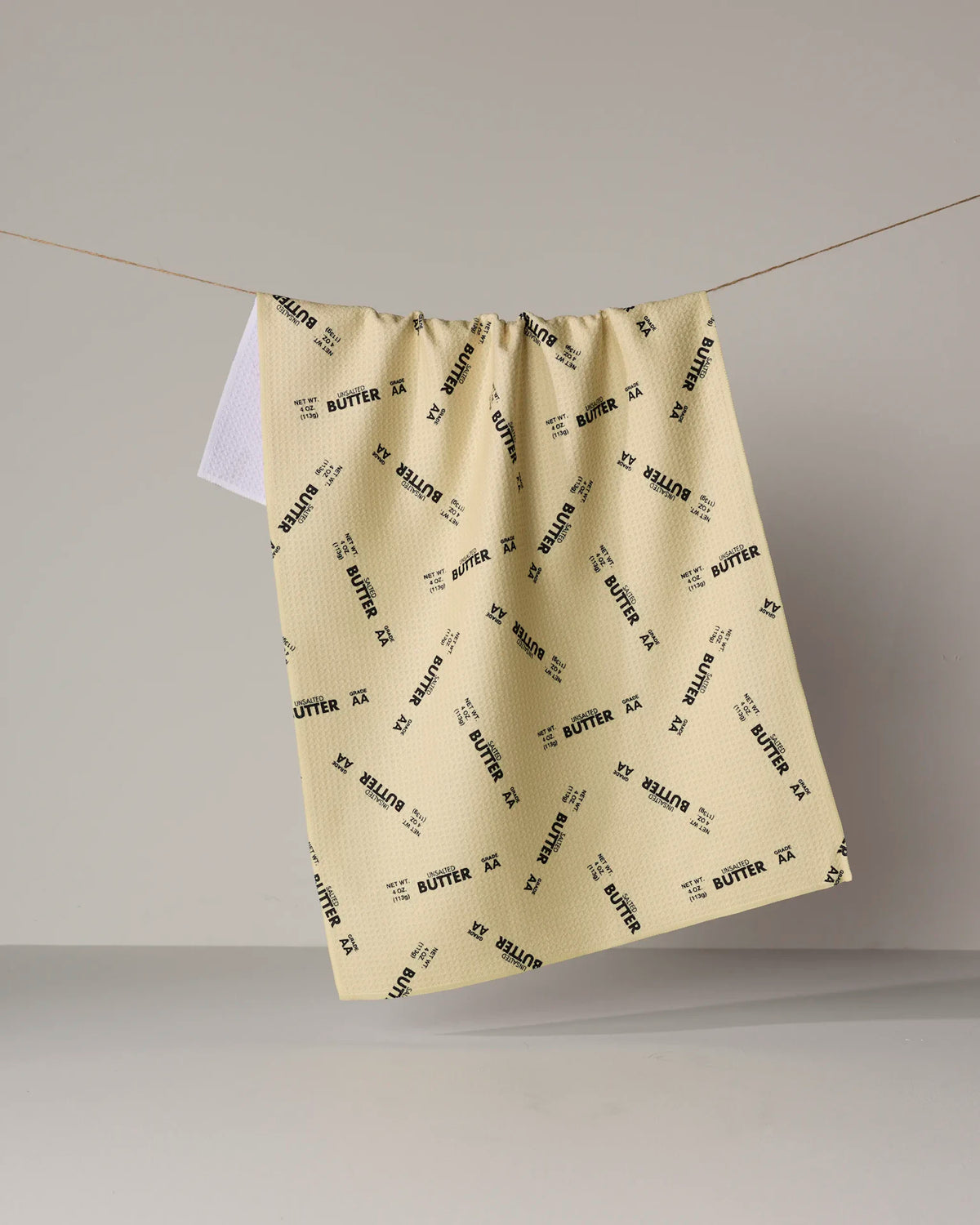 A cream-colored, quick-drying GEOMETRY Salted vs Unsalted kitchen towel hanging on a clothesline with a repeated pattern of butter wrapper graphics.