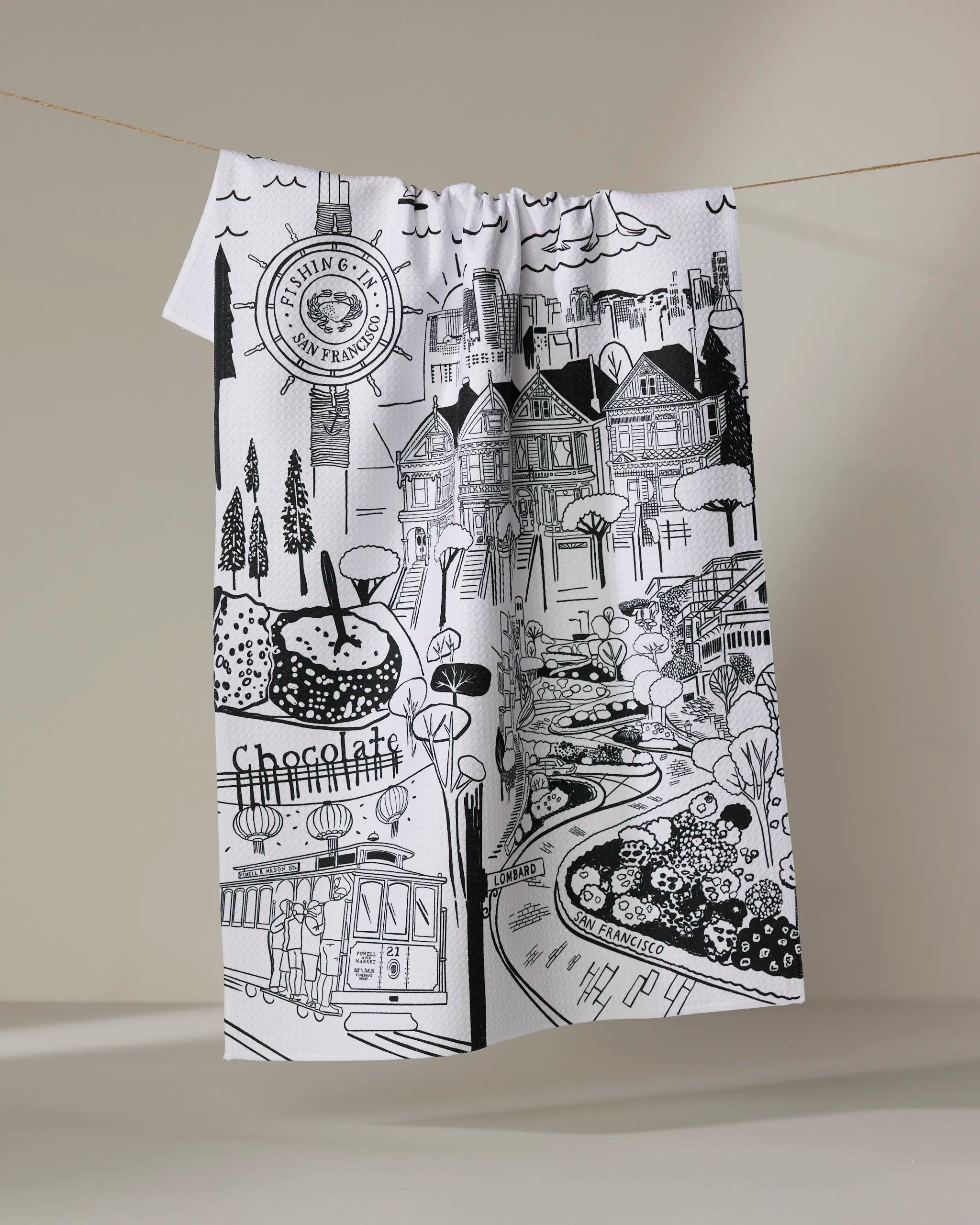 A white recycled kitchen towel by Elisabeth Wing, featuring black line drawings of San Francisco landmarks and the word "chocolate," hangs on a clothesline against a plain background.