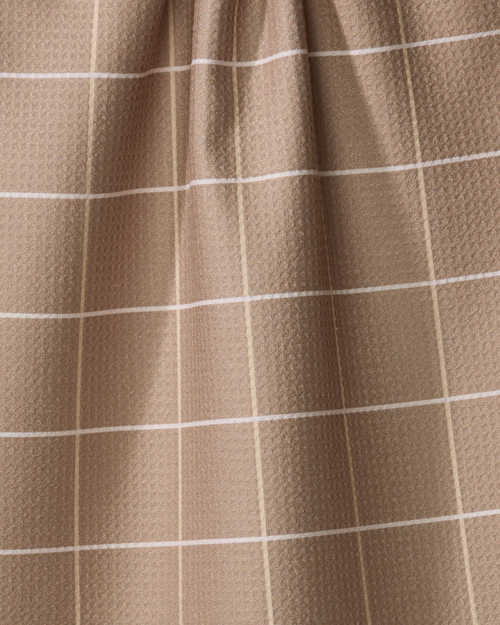 The Sand Grid kitchen towel by Kelsi Fullmer features a light brown recycled fabric with white and beige grid lines forming squares and a subtle textured pattern. The top is slightly gathered, creating gentle folds.