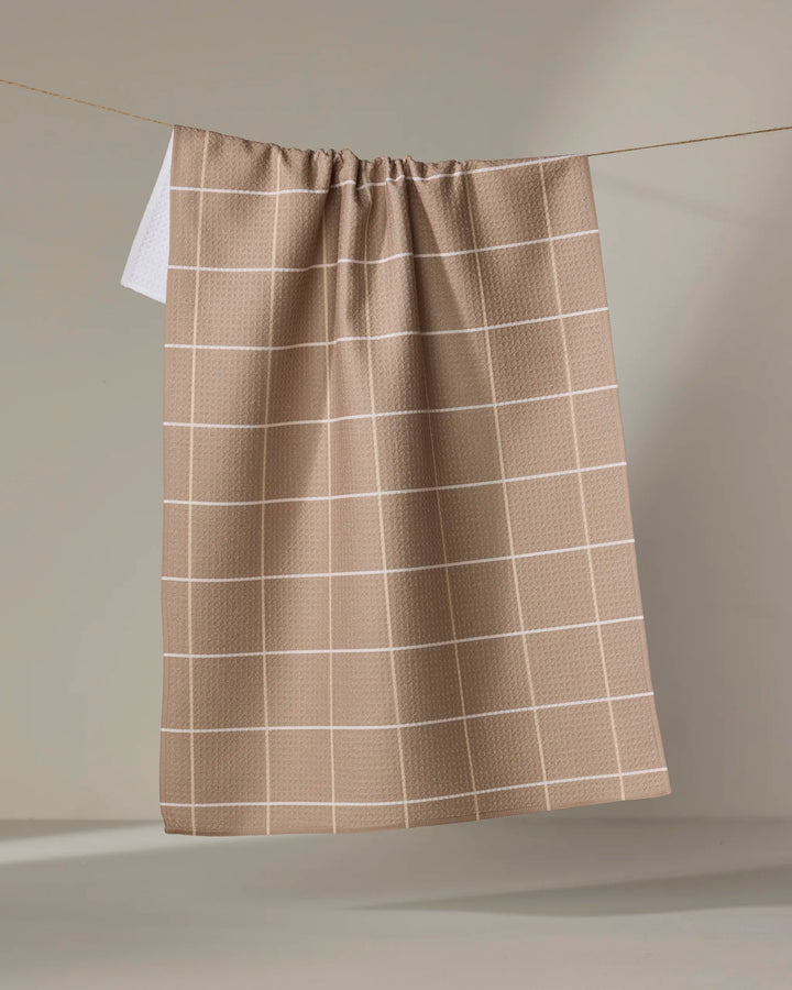 The "Sand Grid" recycled kitchen towel by Kelsi Fullmer, featuring a white grid pattern in beige, hangs on a line against a neutral background.