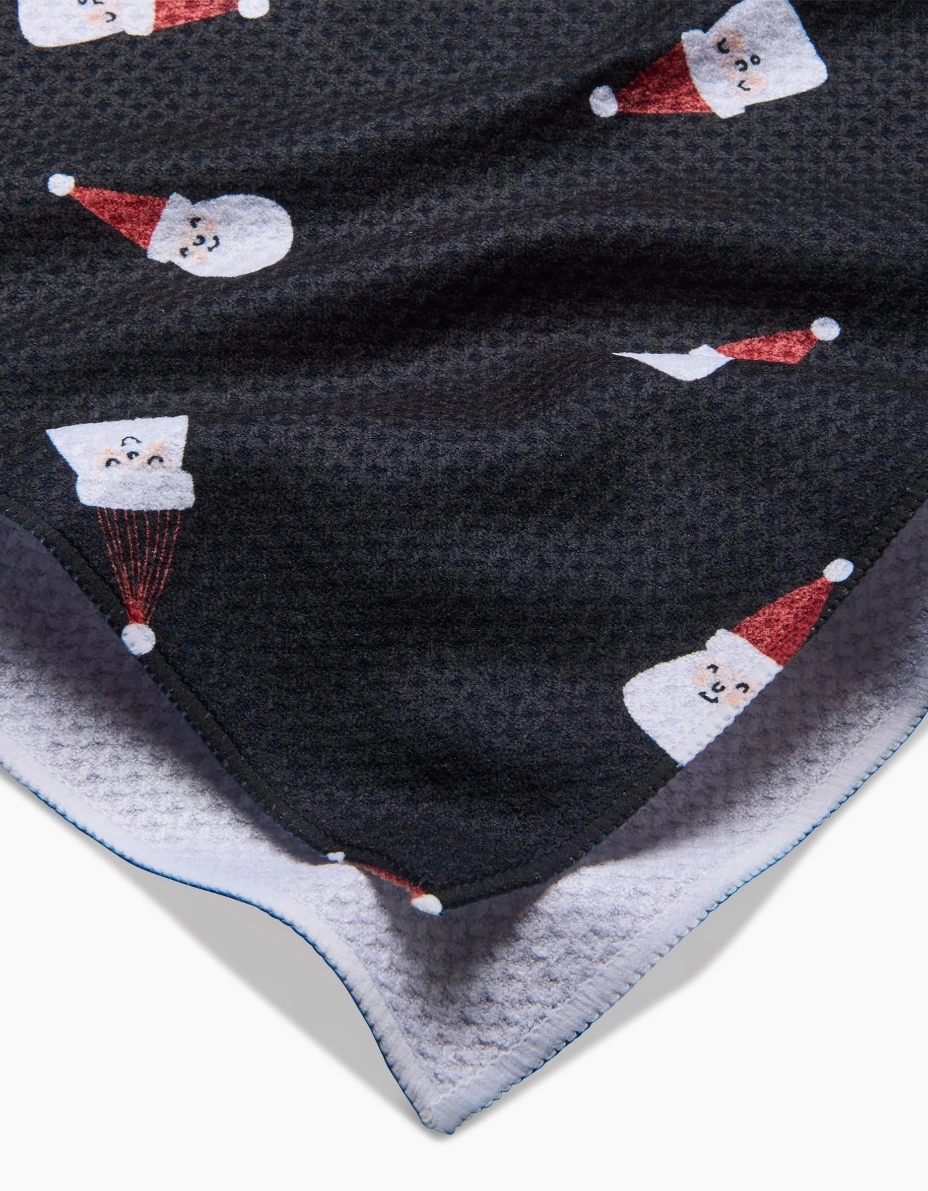 A close-up of the Coit Creative Santa Faces tea towel, featuring a textured black fabric with smiling Santas in red hats, layered over a white, waffle-textured recycled kitchen towel.