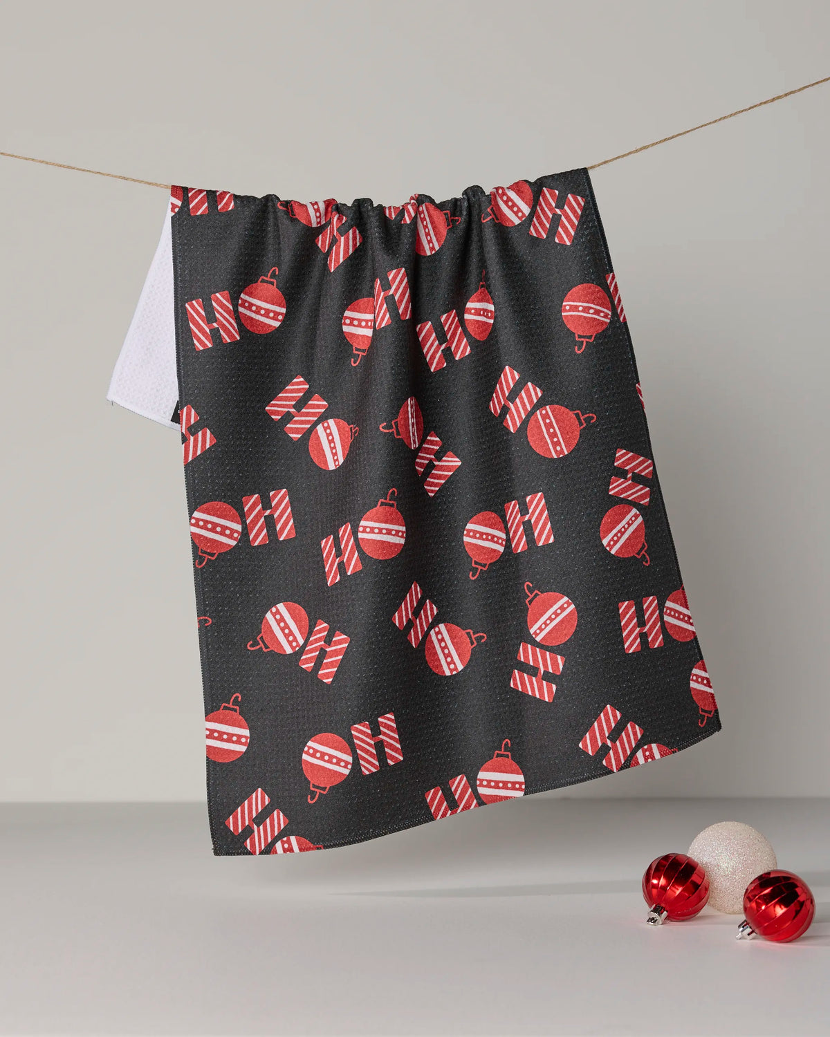 A Geometry “Santa Greetings” black recycled kitchen towel featuring red and white candy cane and ornament patterns hangs on a line, while two red and white ornaments rest on the light surface below.