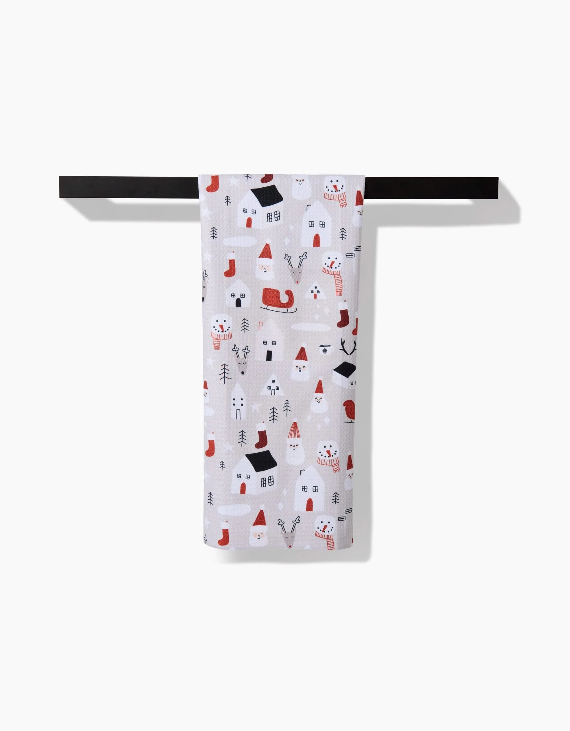 The Coit Creative Santas Village kitchen tea towel, featuring Santa faces, reindeer, houses, and Christmas trees in red, black, and gray, hangs on a black bar against a white background.
